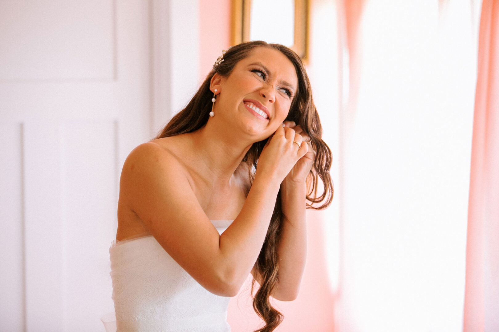 A woman in a white strapless dress adjusts her earring, smiling softly. She stands near a window with pink curtains, the atmosphere buzzing with excitement for the Willow Creek Winery wedding.