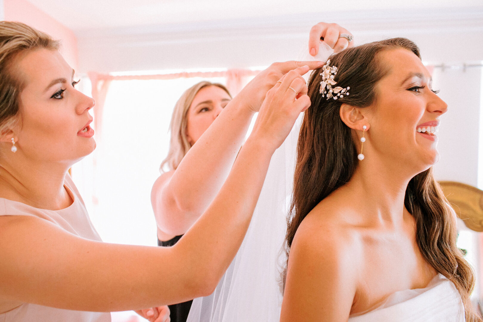 In a brightly lit room at Willow Creek Winery, two women assist a smiling bride with her hair accessories and veil.