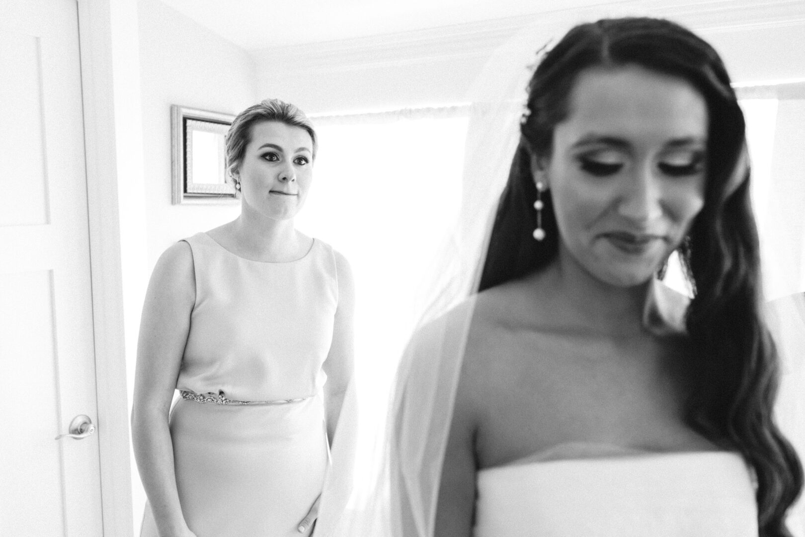 In a sunlit room, the bride in her elegant wedding dress beams with joy at her Willow Creek Winery wedding, while another woman in a light dress stands behind her, admiring the moment.