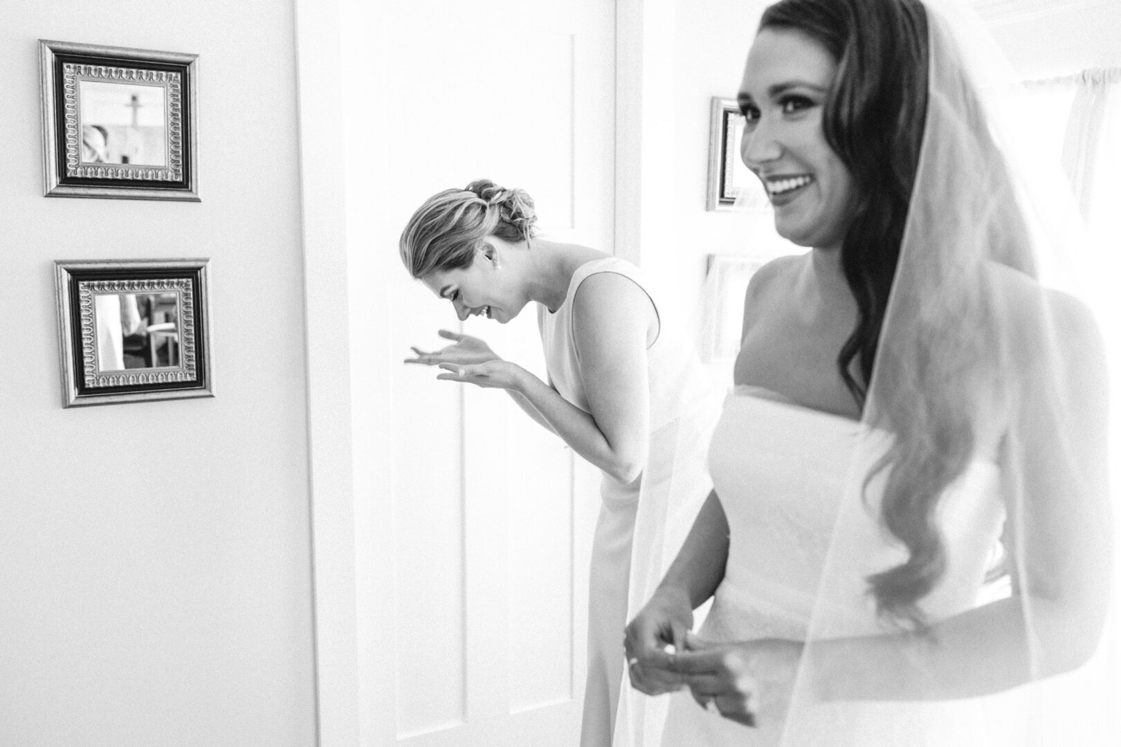 A bride and her bridesmaid share a laugh in a sunlit room at Willow Creek Winery. The bride, in her strapless dress and veil, and the bridesmaid, in a sleeveless gown, stand near framed photos on the wall, capturing moments of this joyful wedding day.