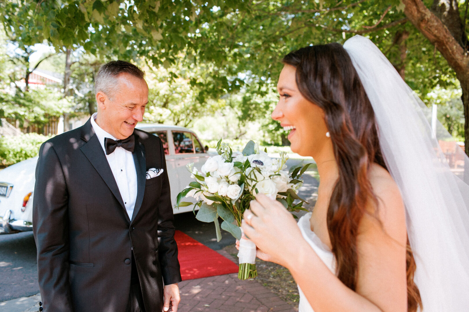 A bride holding a bouquet smiles at a man in a tuxedo, the enchanting setting of Willow Creek Winery providing the perfect backdrop with its white car and red carpet under elegant trees.