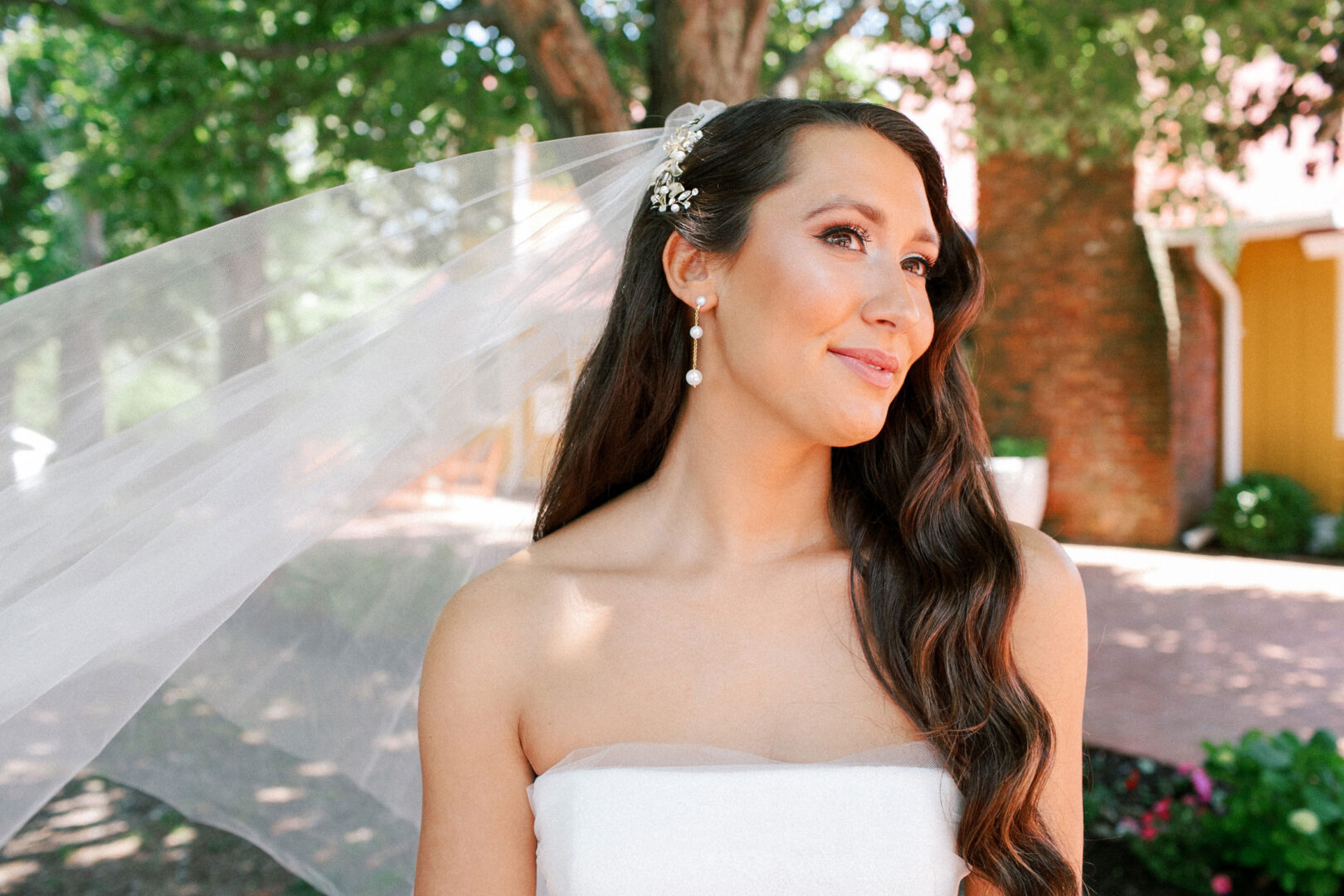 At a picturesque Willow Creek Winery wedding, a bride with long hair and a veil stands outdoors, elegantly adorned in a strapless white dress and pearl earrings.