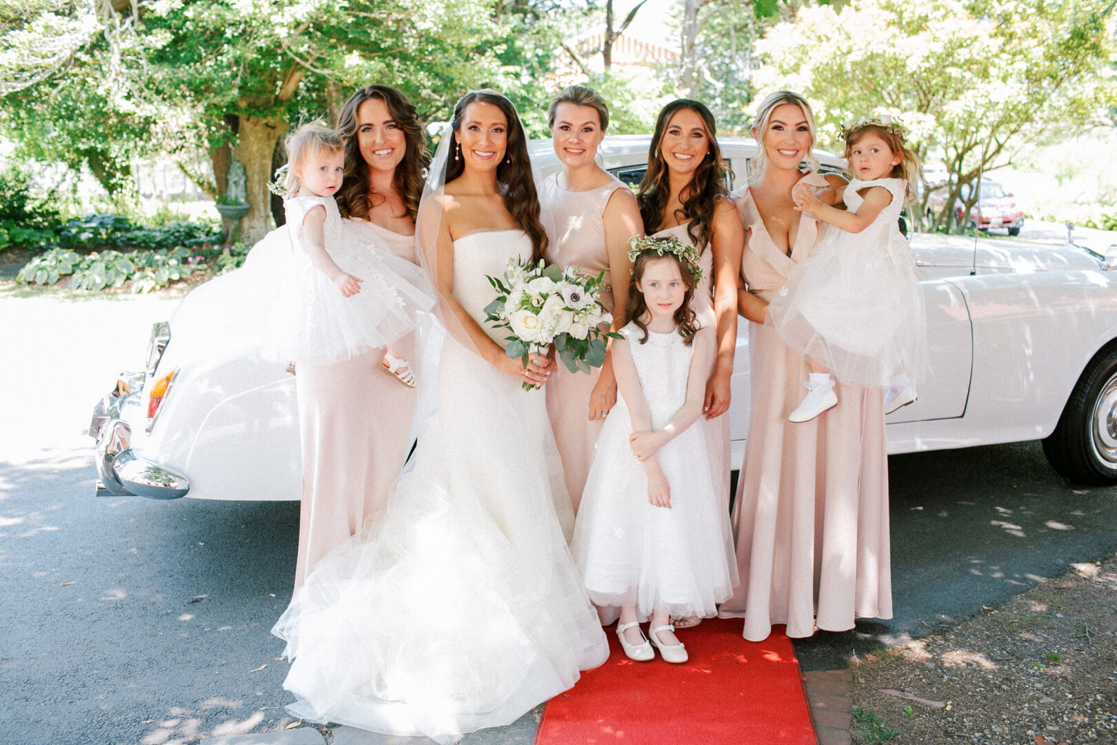 Bride and bridesmaids in elegant beige dresses stand on a red carpet alongside two young girls in white dresses, all set against the charming backdrop of a vintage car at the enchanting Willow Creek Winery wedding.