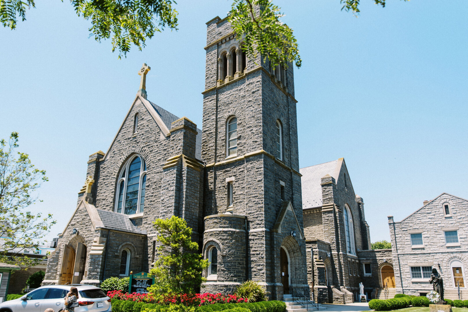 The stone church, reminiscent of a charming Willow Creek Winery wedding, features arched windows and a towering bell with an elegant cross atop. Nestled among lush greenery and vibrant flowers, the scene is complemented by a car parked serenely under the clear blue sky.