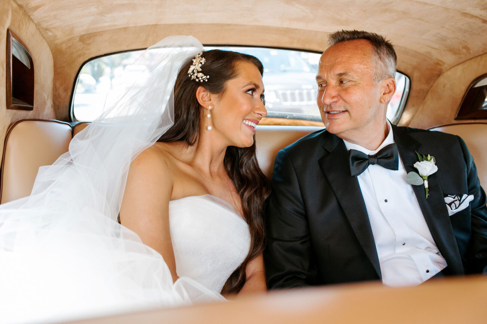 A bride and groom sit together in the backseat of a car, dressed in a wedding gown and tuxedo, looking at each other and smiling, fresh from their beautiful Willow Creek Winery wedding.