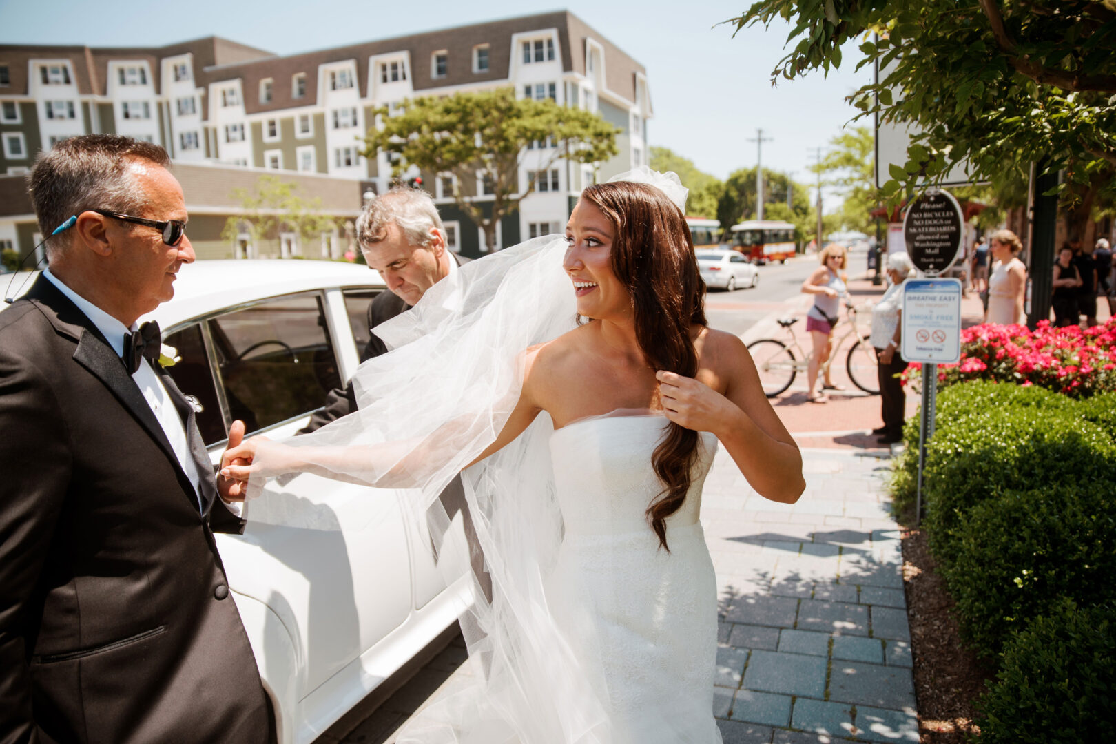 Bride in white dress with veil steps out of a car, assisted by two men in suits, surrounded by the lush greenery and rustic charm of Willow Creek Winery wedding.