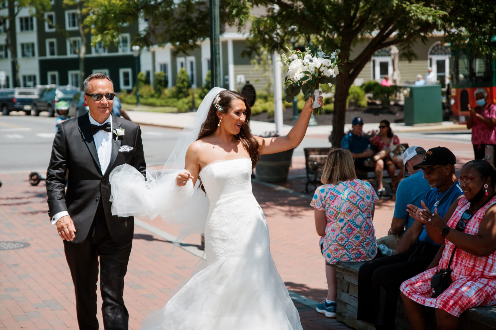 At the Willow Creek Winery wedding, a bride in a white dress and veil walks with a man in a suit. She holds up her bouquet, smiling as people sit and clap nearby, with trees and buildings in the background.