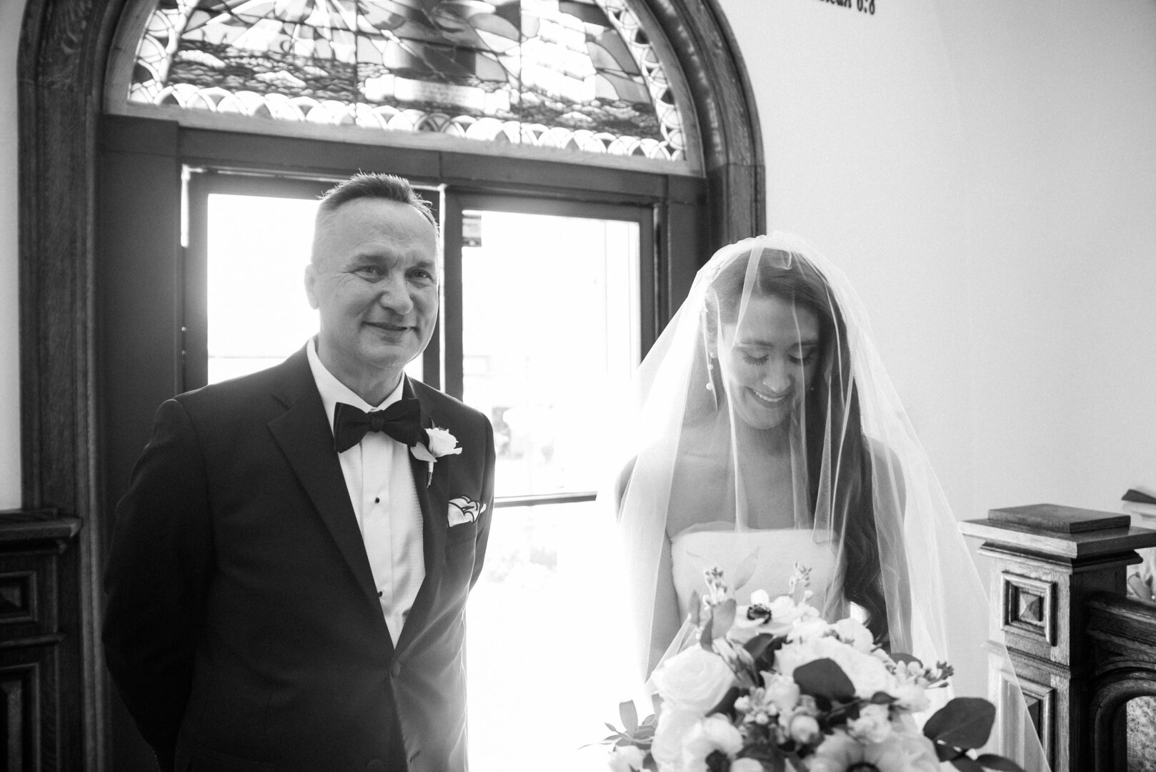 A bride in a veil holding a bouquet stands beside a man in a suit inside Willow Creek Winery, their silhouettes illuminated by the vibrant stained glass window.