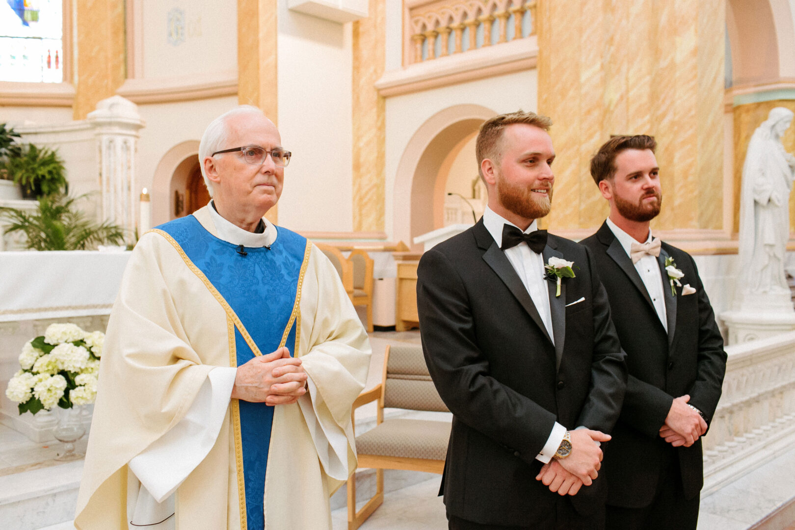 A priest in white and blue robes stands beside two groomsmen in black tuxedos with bow ties inside the church, setting the stage for a Willow Creek Winery wedding.