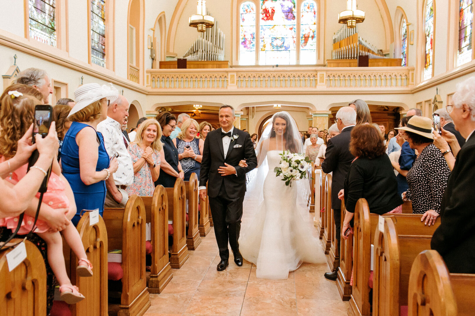 A bride and groom walk down the aisle at Willow Creek Winery, surrounded by seated guests and vibrant stained glass windows.