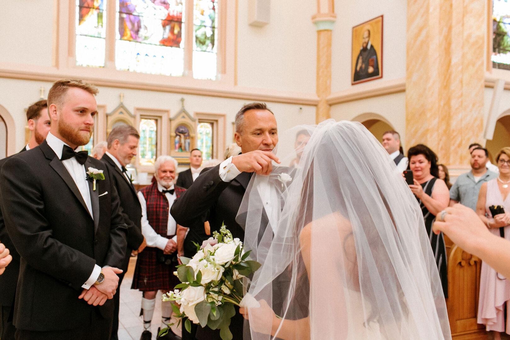 A bride in a veil stands in the serene atmosphere of Willow Creek Winery. A man adjusts her veil while the groom and guests look on, surrounded by stained glass windows and religious icons that add to the sacred ambiance of this special day.