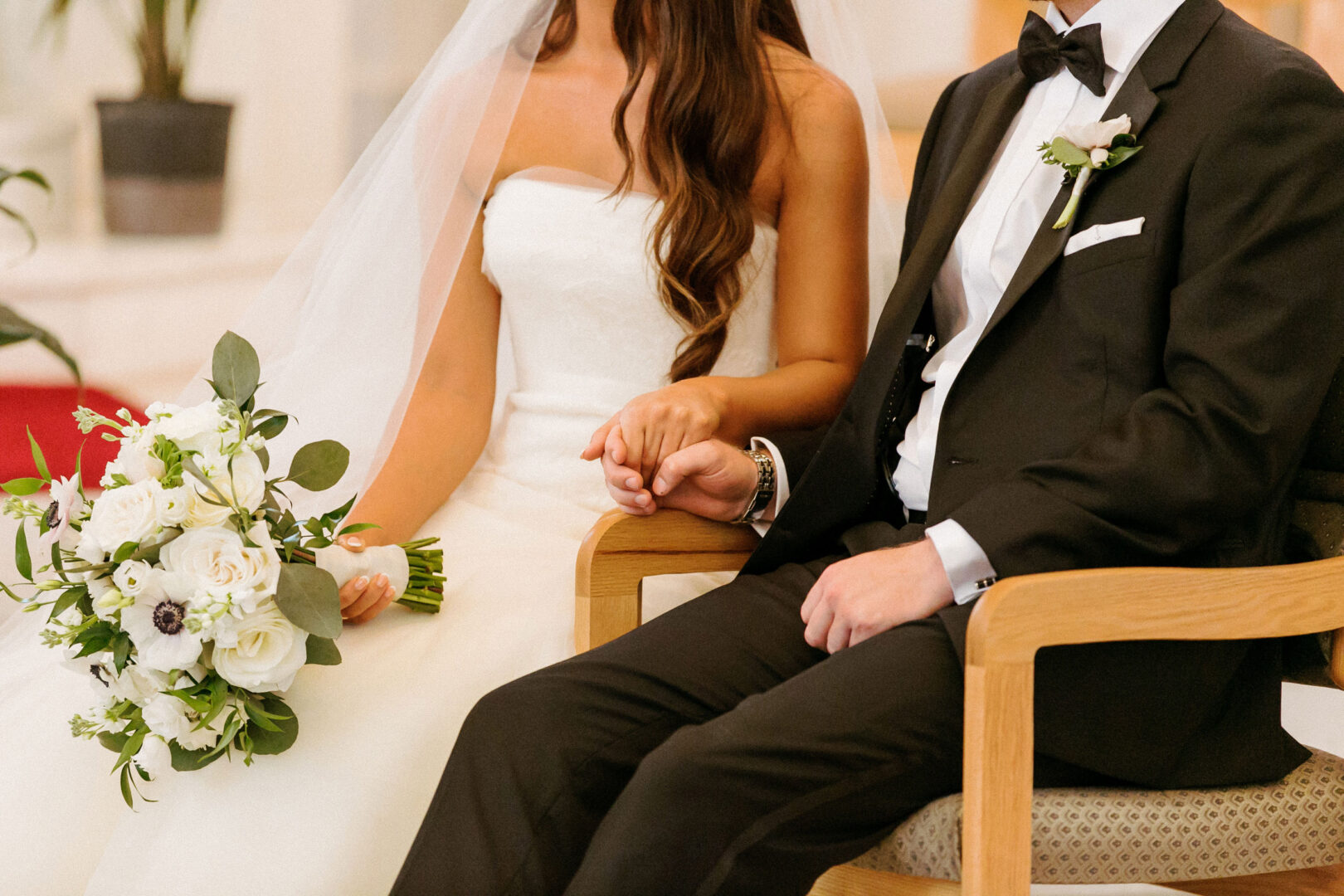 At their Willow Creek Winery wedding, the bride and groom sit holding hands. The bride, in a white strapless gown and veil, holds a bouquet of white flowers, while the groom complements her in a black suit with a bow tie.