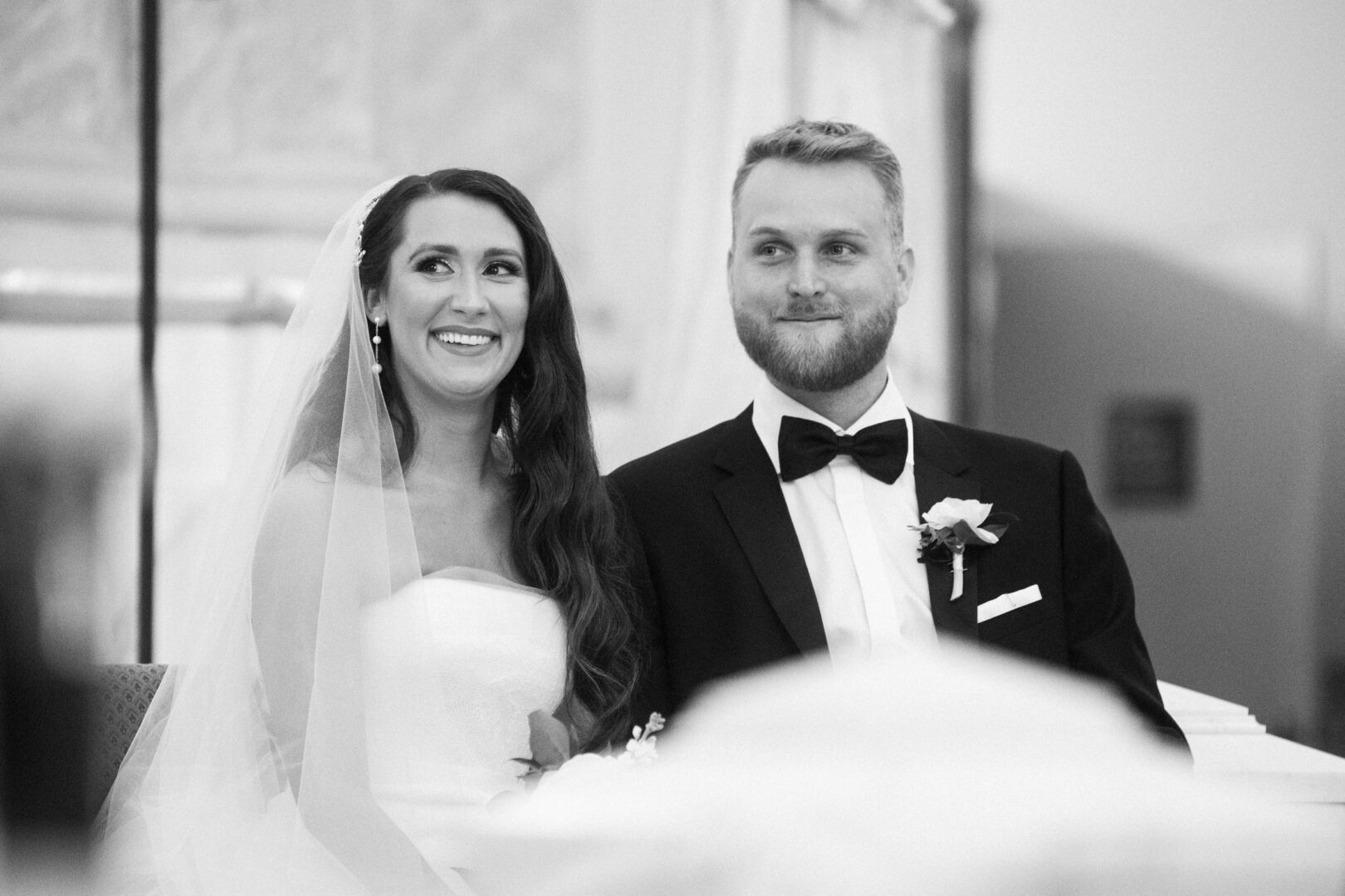 At a Willow Creek Winery wedding, a bride and groom sit together, smiling during the ceremony. The bride wears a veil and strapless dress, while the groom is in a tuxedo.