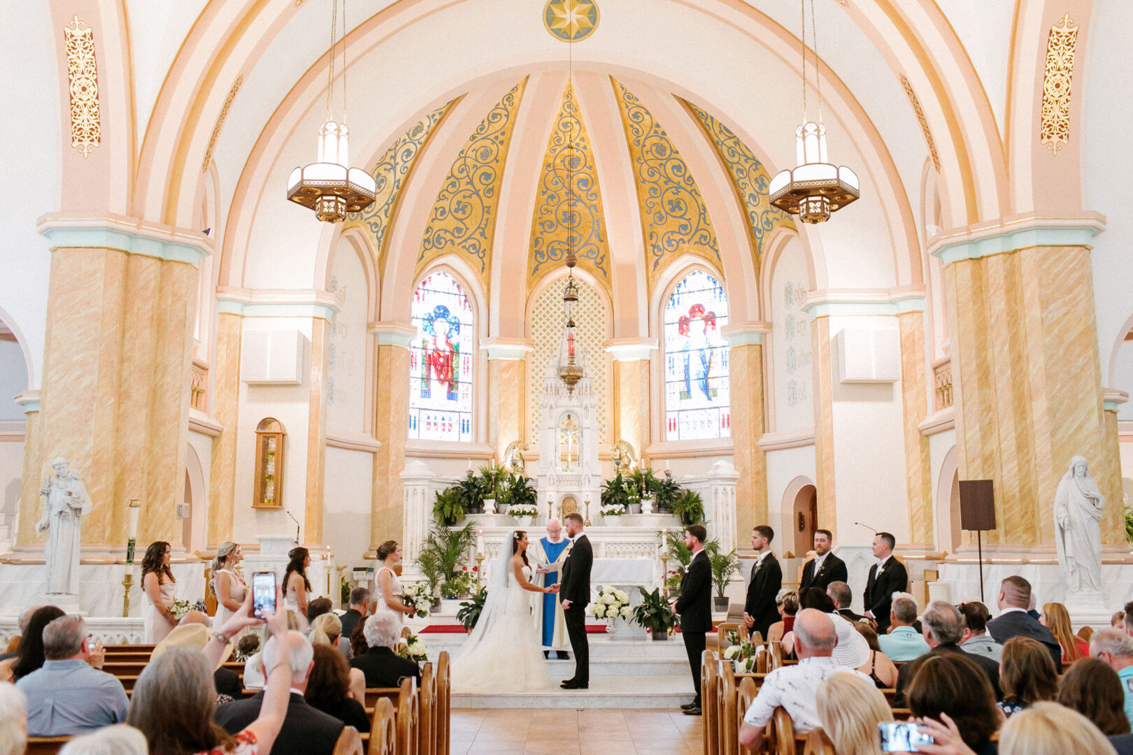 A couple stands at the altar during a Willow Creek Winery wedding ceremony in a church with stained glass windows. Guests are seated, and some are taking photos.