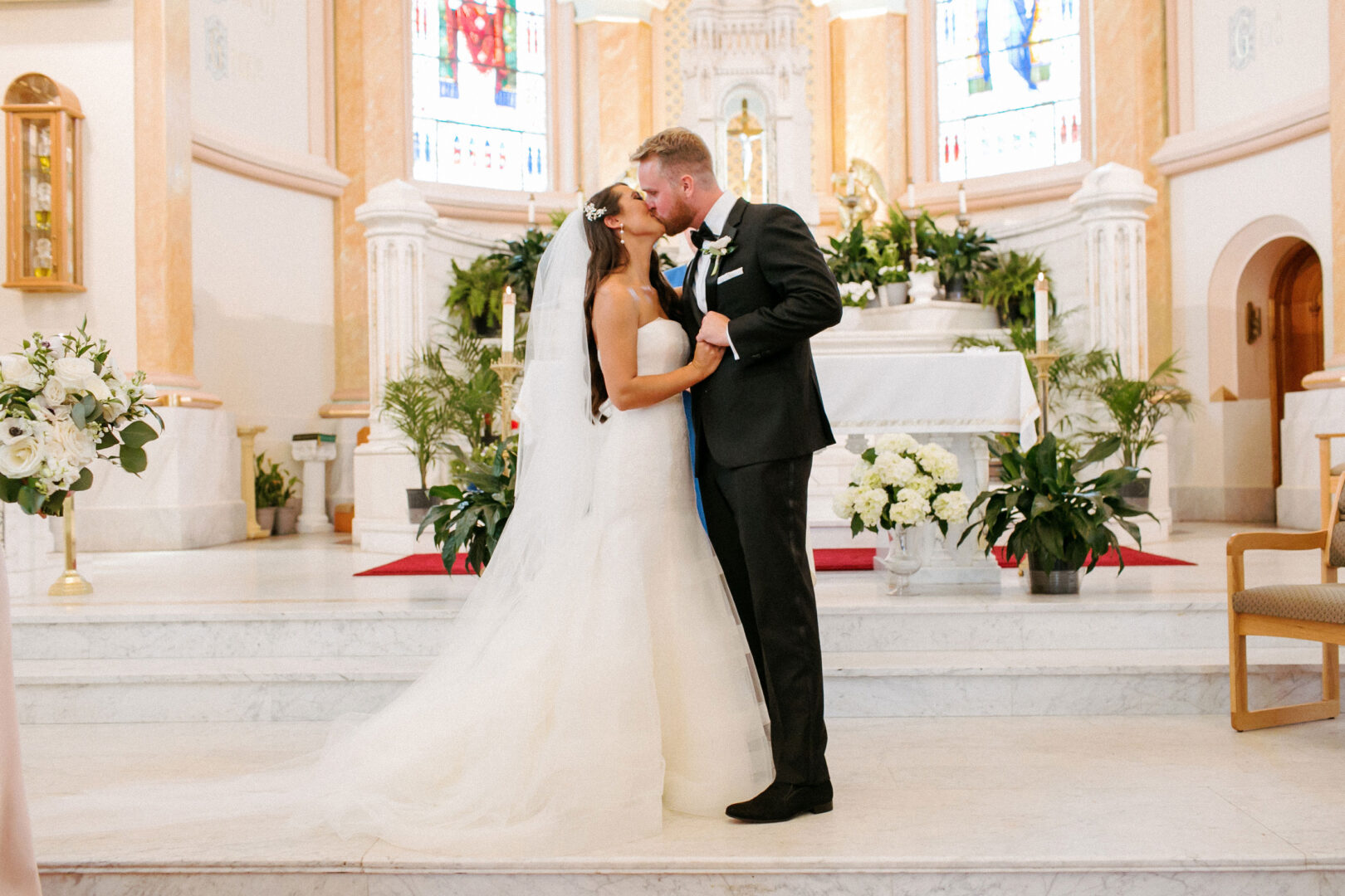 A newlywed couple shares a kiss at the altar in a church adorned with flowers and greenery, their journey beginning just like the serene romance of a Willow Creek Winery wedding.