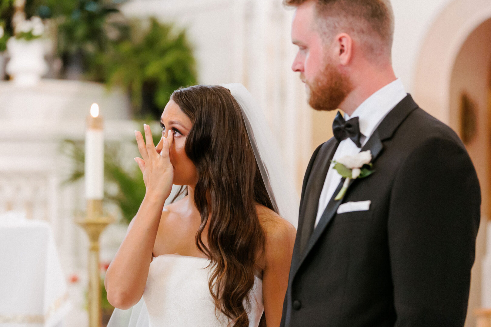 At a Willow Creek Winery wedding, a bride wipes her tears during the ceremony, standing next to a groom in a black suit and white boutonniere.