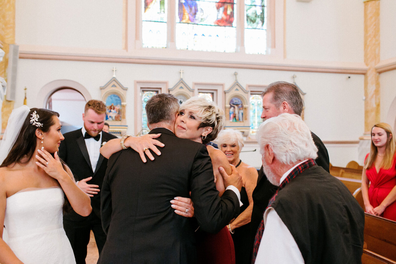 At a quaint Willow Creek Winery wedding, a bride and groom in the church share a cherished moment with family members, including a woman embracing an older man, surrounded by joyous wedding guests.