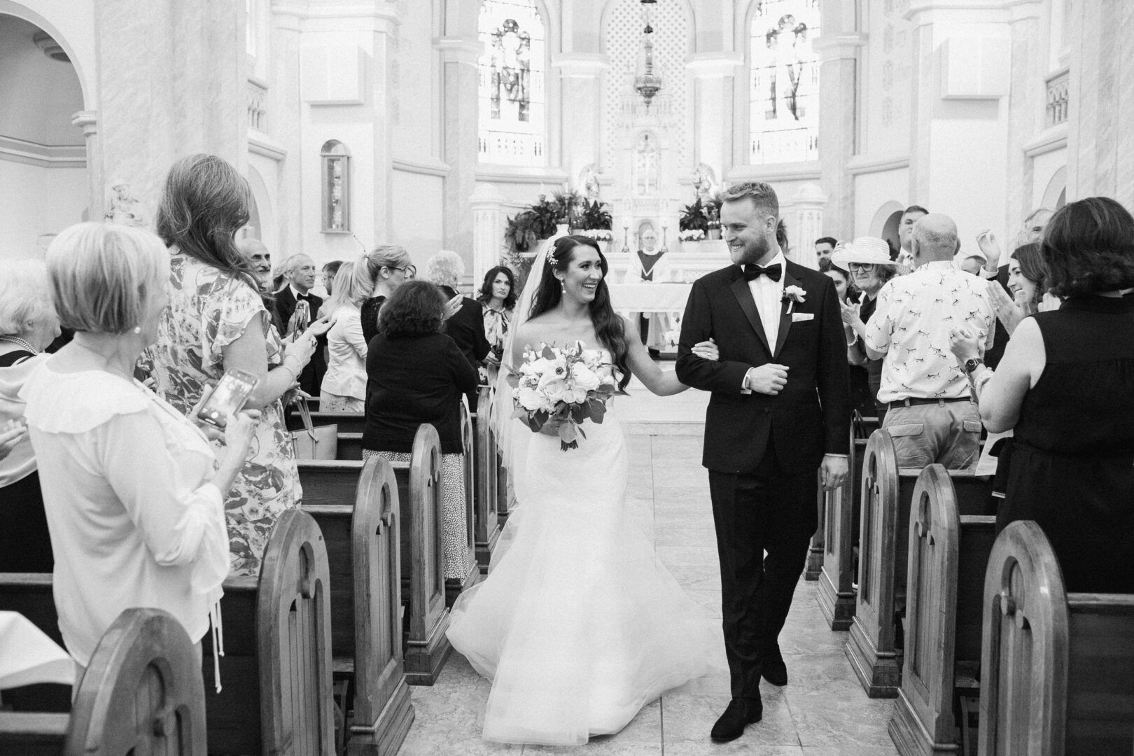 A bride and groom walk down the aisle of Willow Creek Winery, smiling and holding hands, as guests stand and clap in the rustic charm of the venue's chapel.