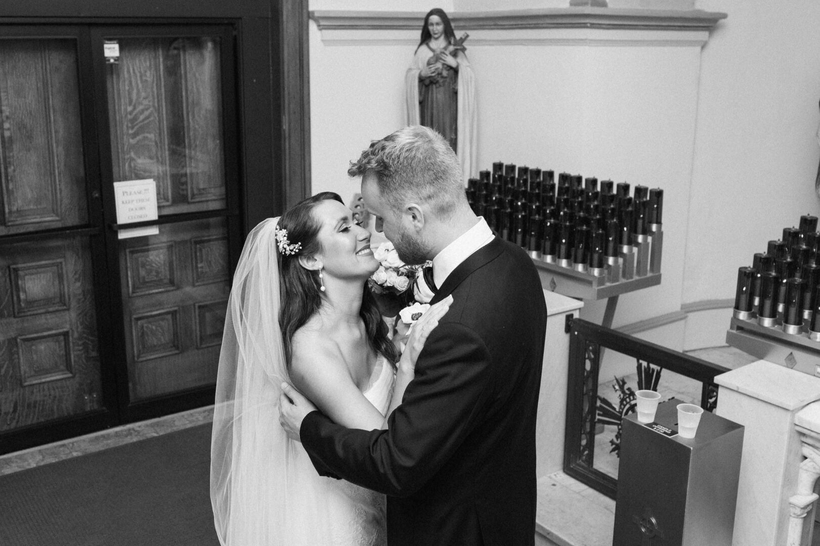 At their Willow Creek Winery wedding, a bride and groom embrace inside a church, framed by the gentle glow of candles and the serene presence of a statue.