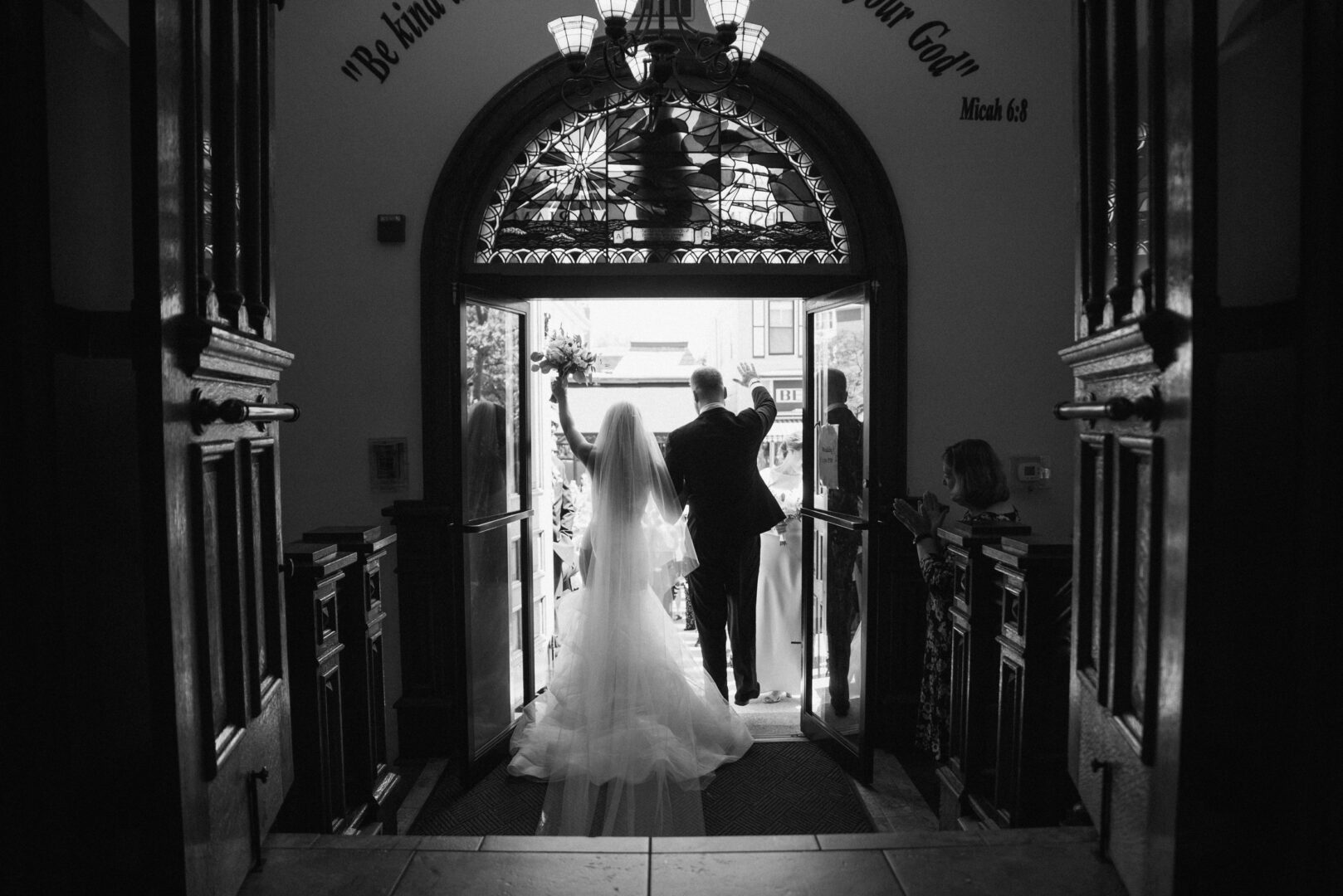 A bride and groom exit a church through large wooden doors, surrounded by ornate stained glass, as sunlight filters in. Guests, who eagerly await the celebration at Willow Creek Winery Wedding, are visible outside in daylight.