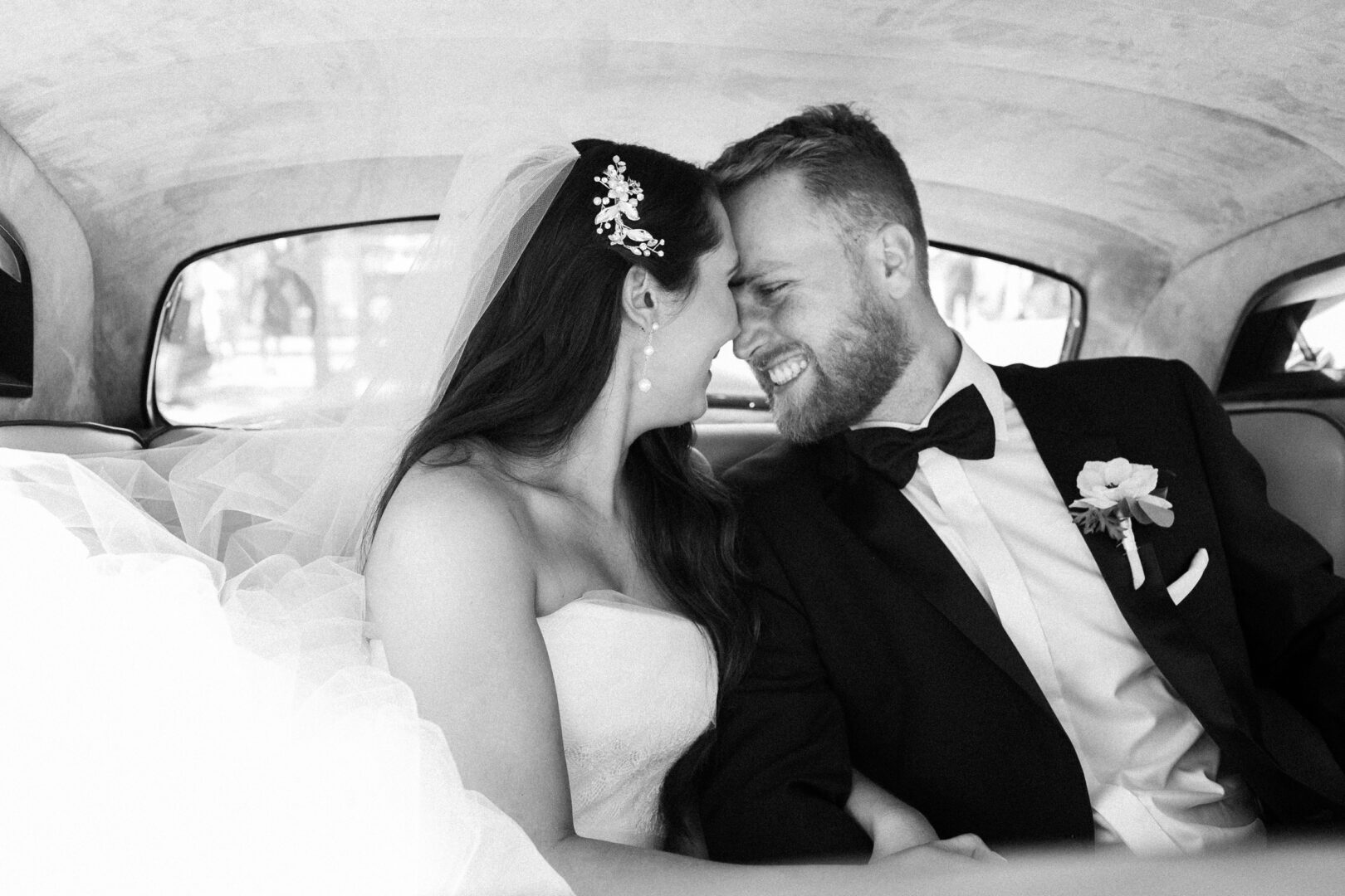 The bride and groom, fresh from their Willow Creek Winery wedding, sit closely together in the backseat of a car, smiling warmly at each other. The bride wears a white gown and veil; the groom is in a black suit with a boutonniere.