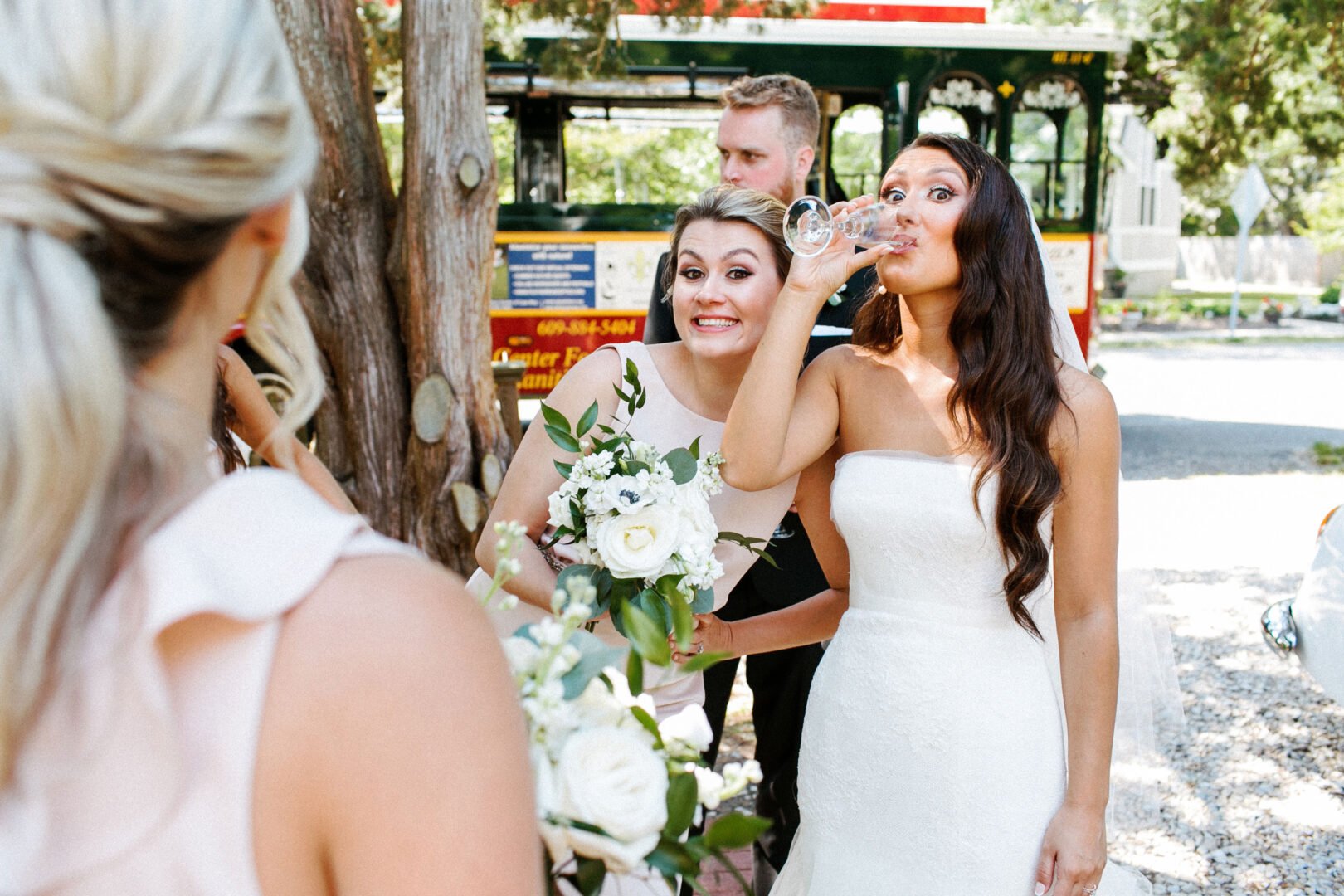 At the elegant Willow Creek Winery wedding, the bride, radiant in her white dress, sips from a glass while holding her bouquet. Beside her, another woman smiles joyfully, capturing the enchanting moment.