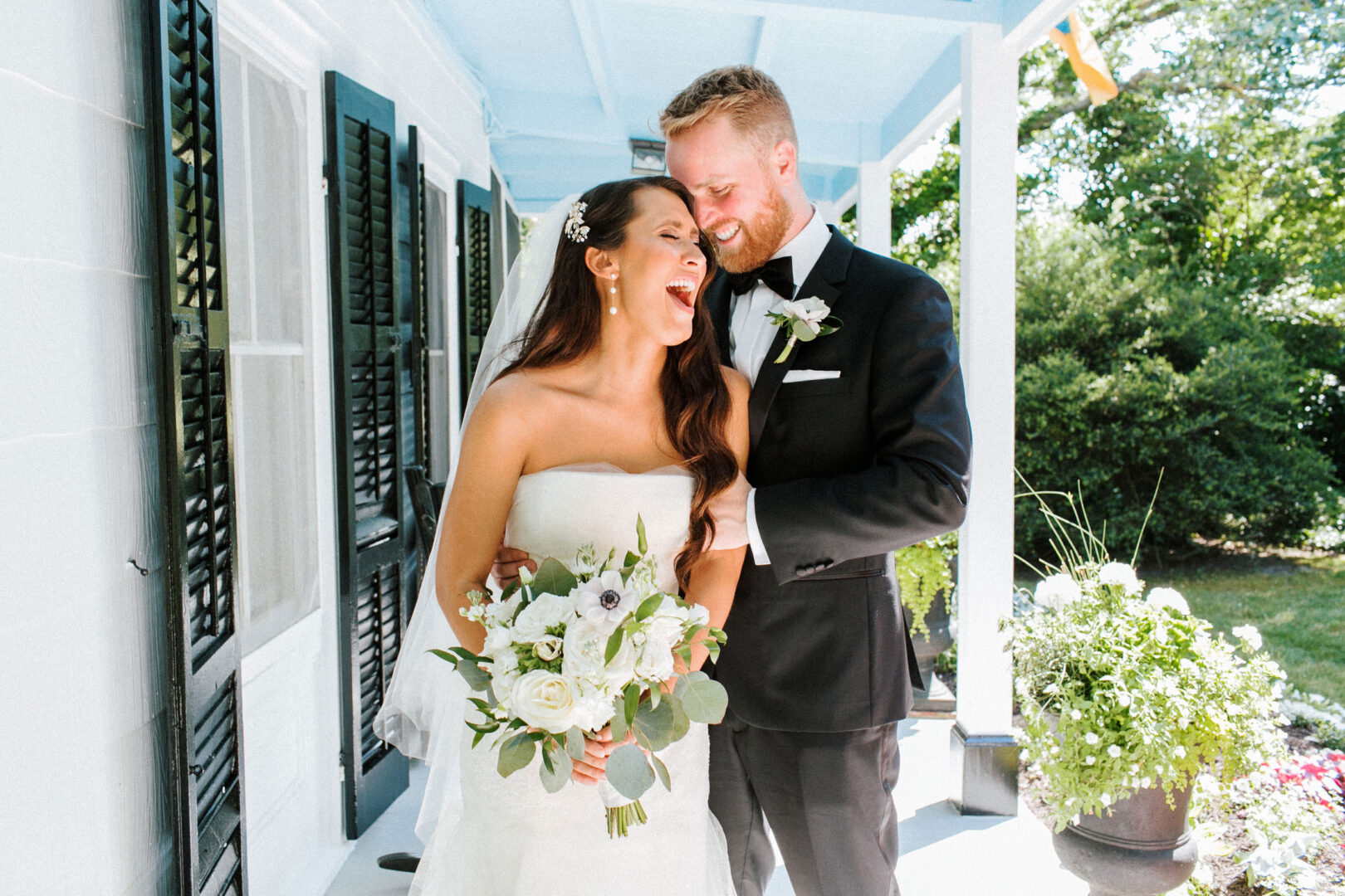 A bride and groom stand together on a porch at Willow Creek Winery, smiling. The bride holds a bouquet of white flowers, perfectly complementing their formal wedding attire.