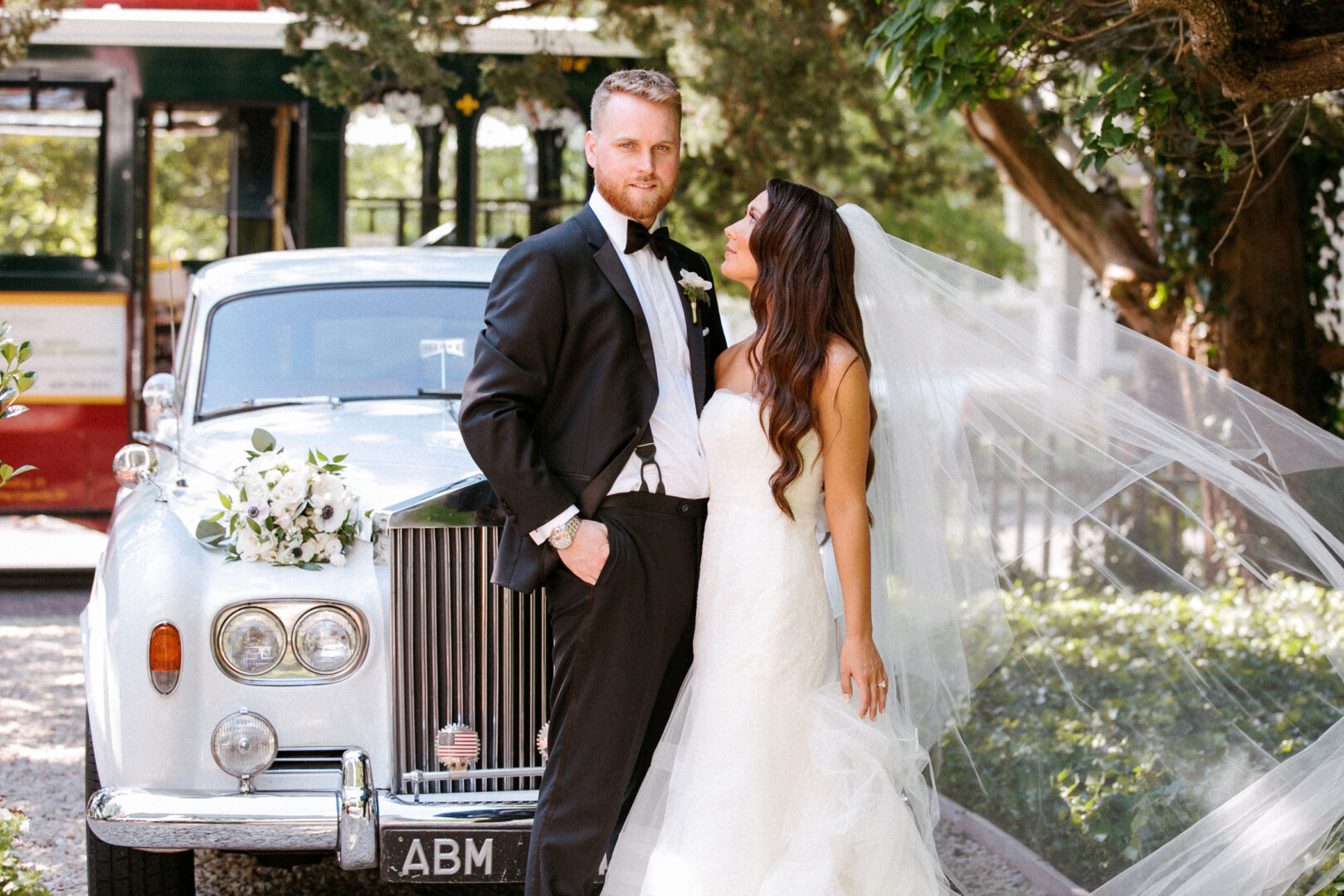 At their Willow Creek Winery wedding, the bride and groom stand beside a classic white car, her veil gracefully flowing in the breeze.