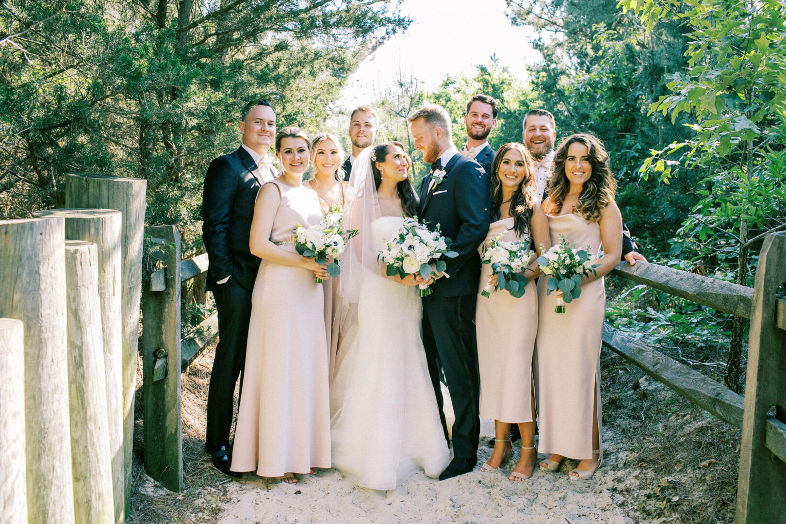 The bride and groom pose with seven members of their wedding party, all dressed in formal attire, surrounded by lush greenery on a sandy path at Willow Creek Winery.