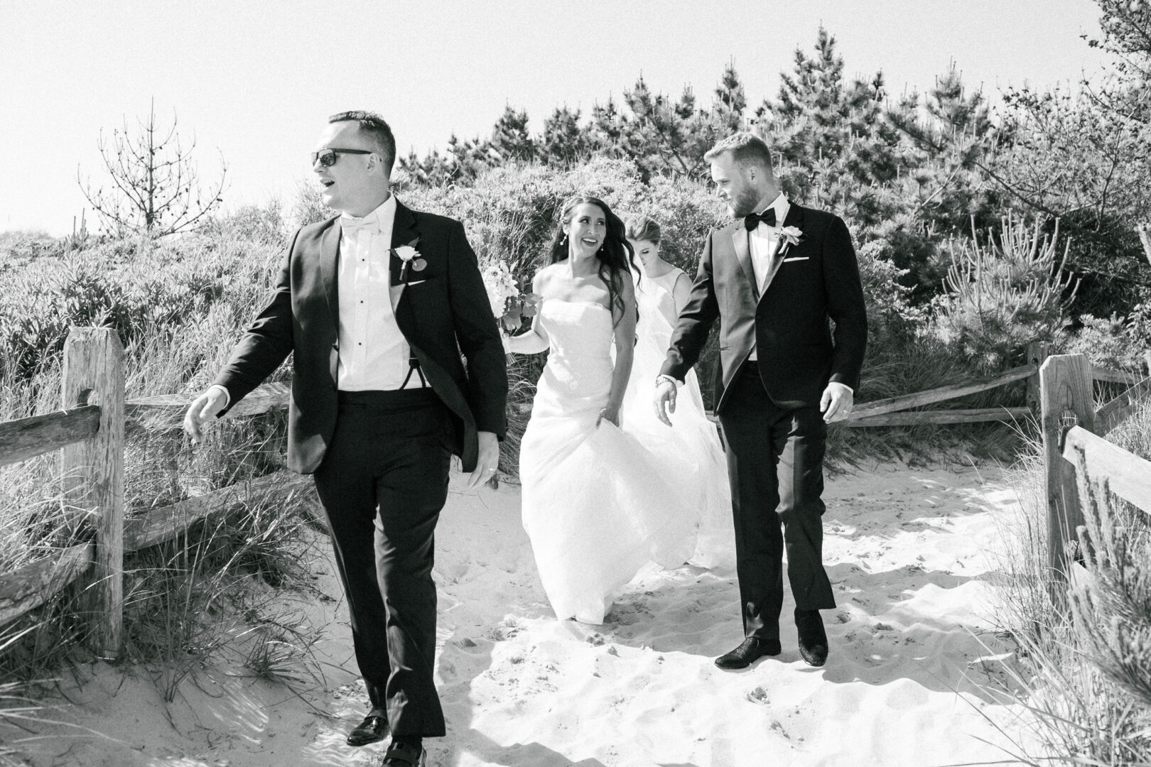 A bride in a white dress strolls along a sandy path at Willow Creek Winery, flanked by two men in tuxedos, with trees gently arching overhead.