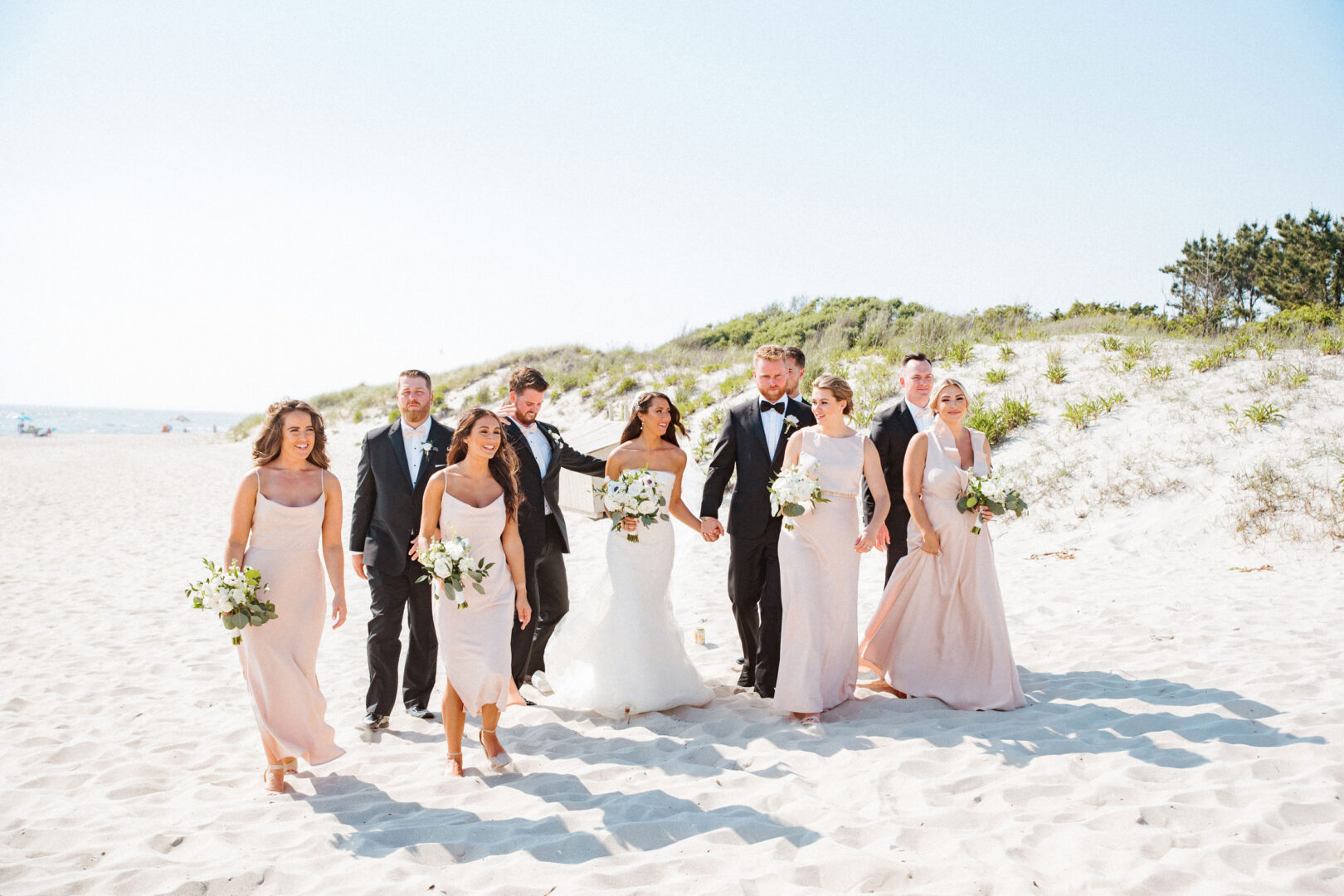 At a picturesque Willow Creek Winery wedding, the party strolls along a sandy beach in formal attire. Bridesmaids dazzle in light pink dresses while groomsmen complement them in dark suits, with the radiant bride and groom taking center stage.