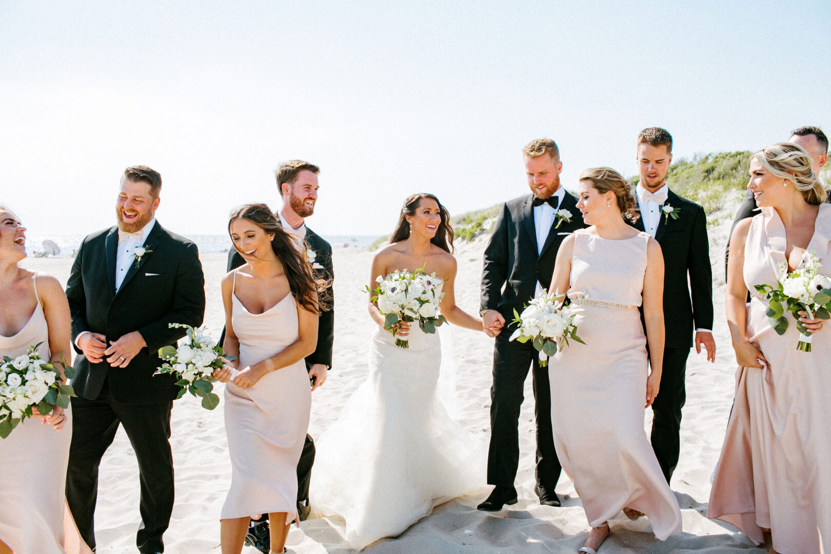 A wedding party strolls along a sandy beach at Willow Creek Winery. The bride and groom are in the center, surrounded by bridesmaids in pale pink dresses and groomsmen in black suits, all holding bouquets of white flowers.