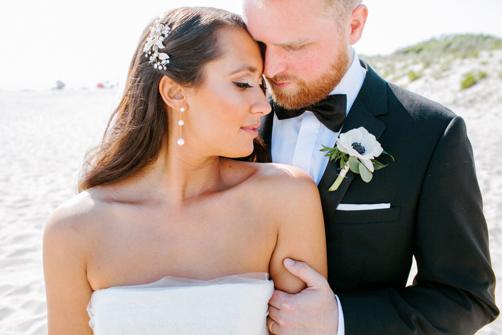 A couple embraces on a sunny beach during their Willow Creek Winery wedding. The groom wears a black suit with a floral boutonniere, while the bride stuns in a strapless dress, jeweled headpiece, and pearl earrings.