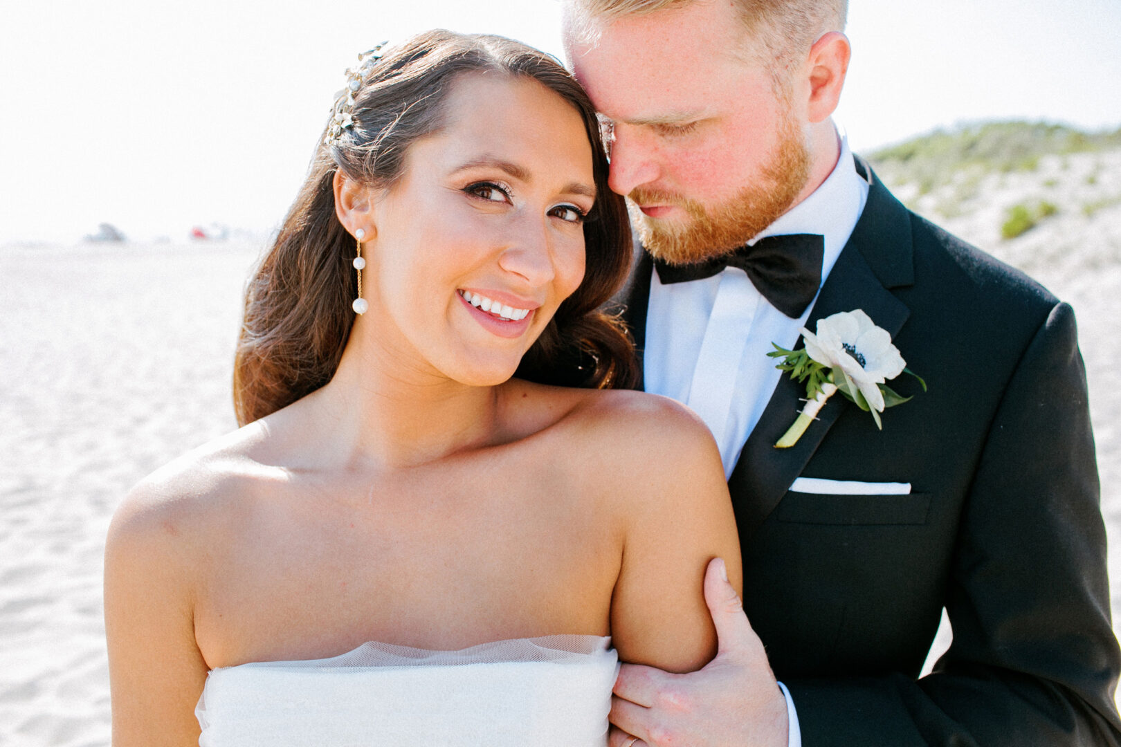 A bride and groom pose on a sunny beach, capturing the joy of their Willow Creek Winery wedding. The bride smiles at the camera, while the groom, in a black suit with a white boutonniere, gazes at her lovingly.