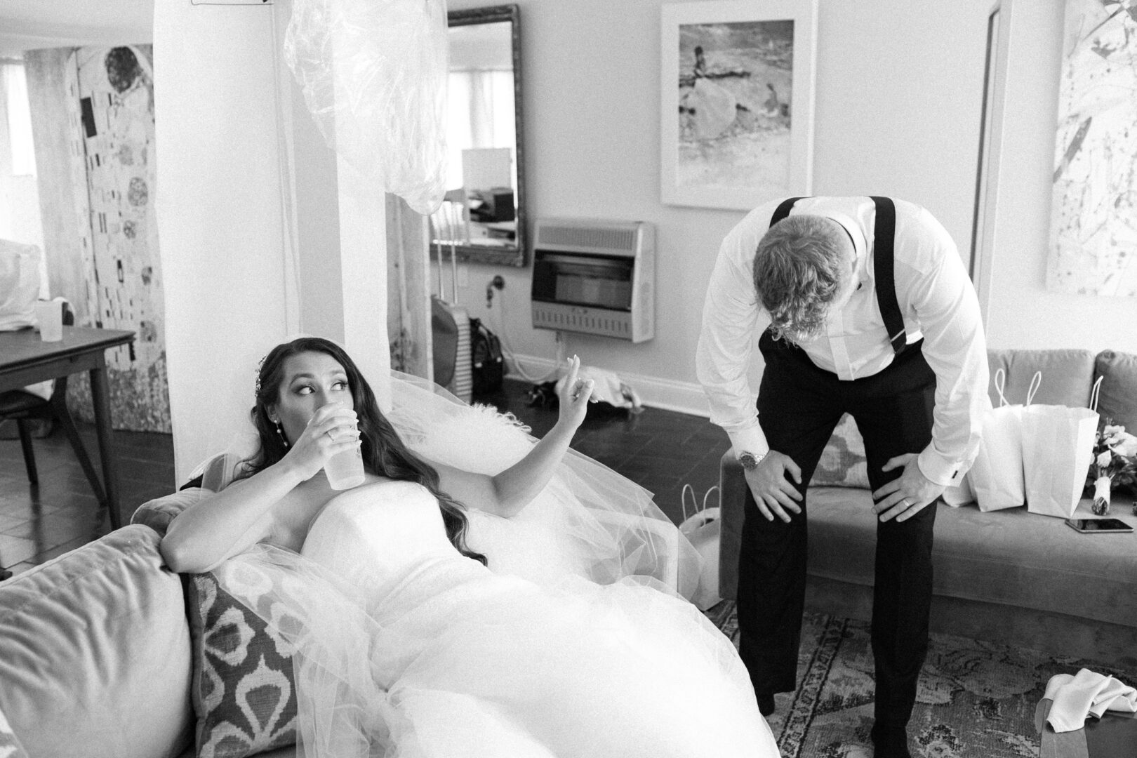 At their Willow Creek Winery wedding, the bride in a stunning dress sits on a couch, sipping from her cup, while the groom in suspenders leans forward beside her.