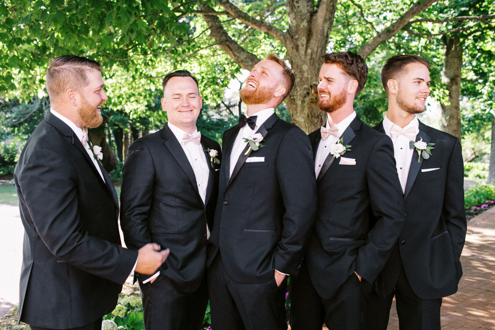 Five men in suits, each wearing a boutonniere, stand together under a tree at Willow Creek Winery, smiling and laughing.