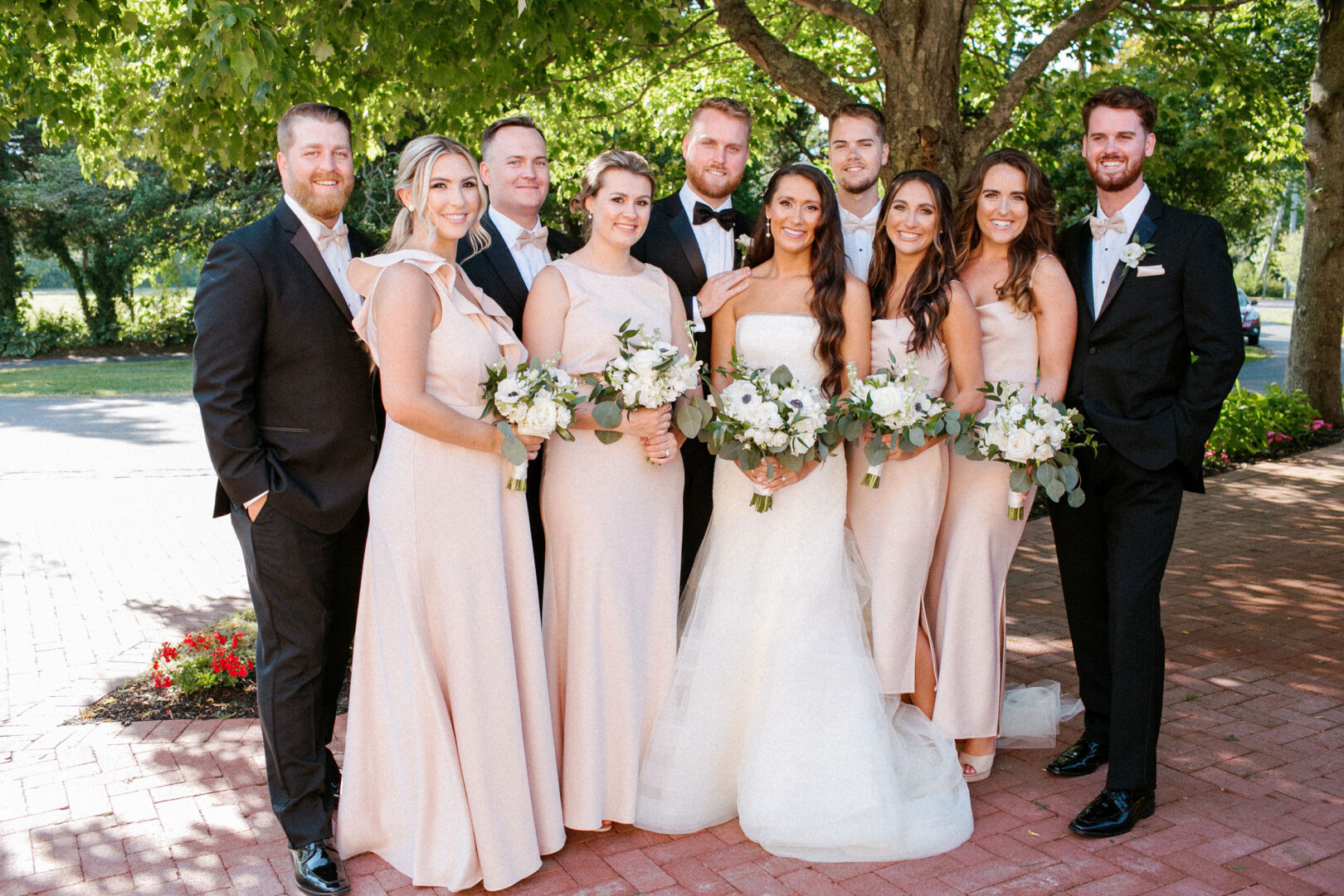 At the picturesque Willow Creek Winery, a wedding party poses outdoors on a charming brick path. Four men in suits and five women in matching light dresses hold bouquets under a large tree, capturing the essence of this enchanting celebration.