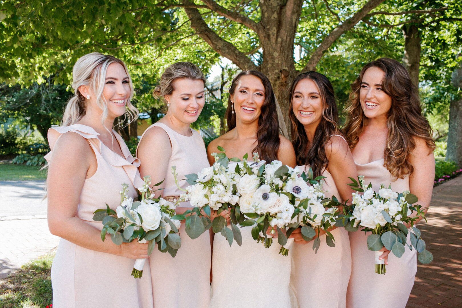 Five women in light dresses stand together outdoors at Willow Creek Winery, smiling and holding bouquets with white flowers and greenery.