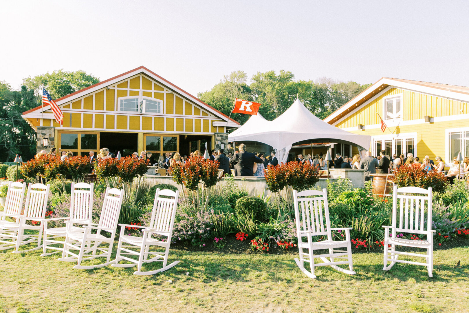 White rocking chairs face two yellow buildings with a tent between them at Willow Creek Winery, perfect for a picturesque wedding. People gather in the area, surrounded by flags and landscaping adorned with vibrant flowers in the foreground.