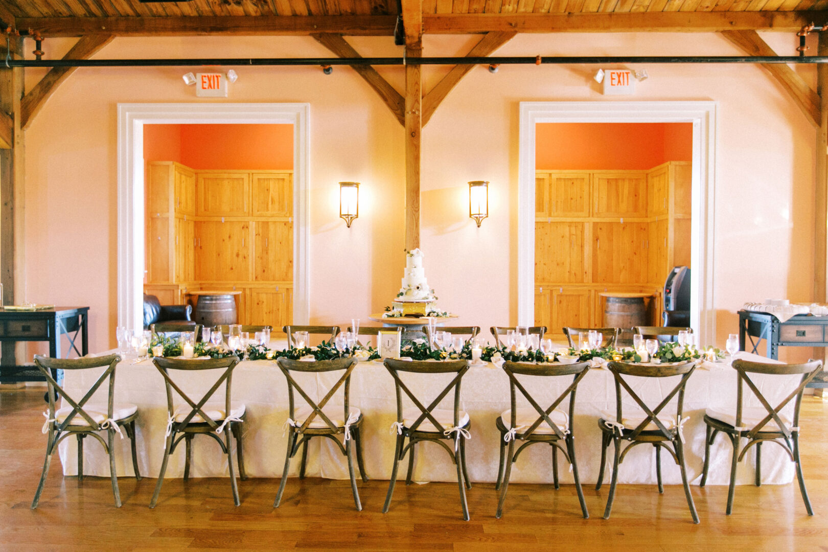 A long dining table set for a formal Willow Creek Winery wedding, with a tiered cake at the center. The room features rustic wooden beams, doors, and exit signs, creating an elegant ambiance for the occasion.