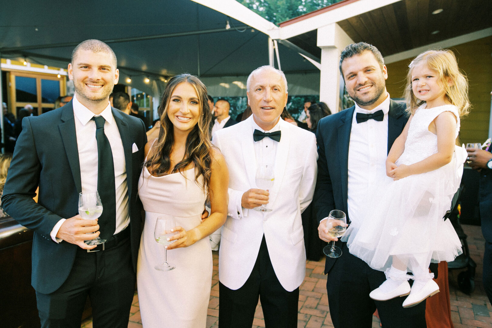 At a Willow Creek Winery wedding, five elegantly dressed adults enjoy their drinks under the sky, accompanied by the joyful presence of a child in a white dress cradled tenderly by one of them.