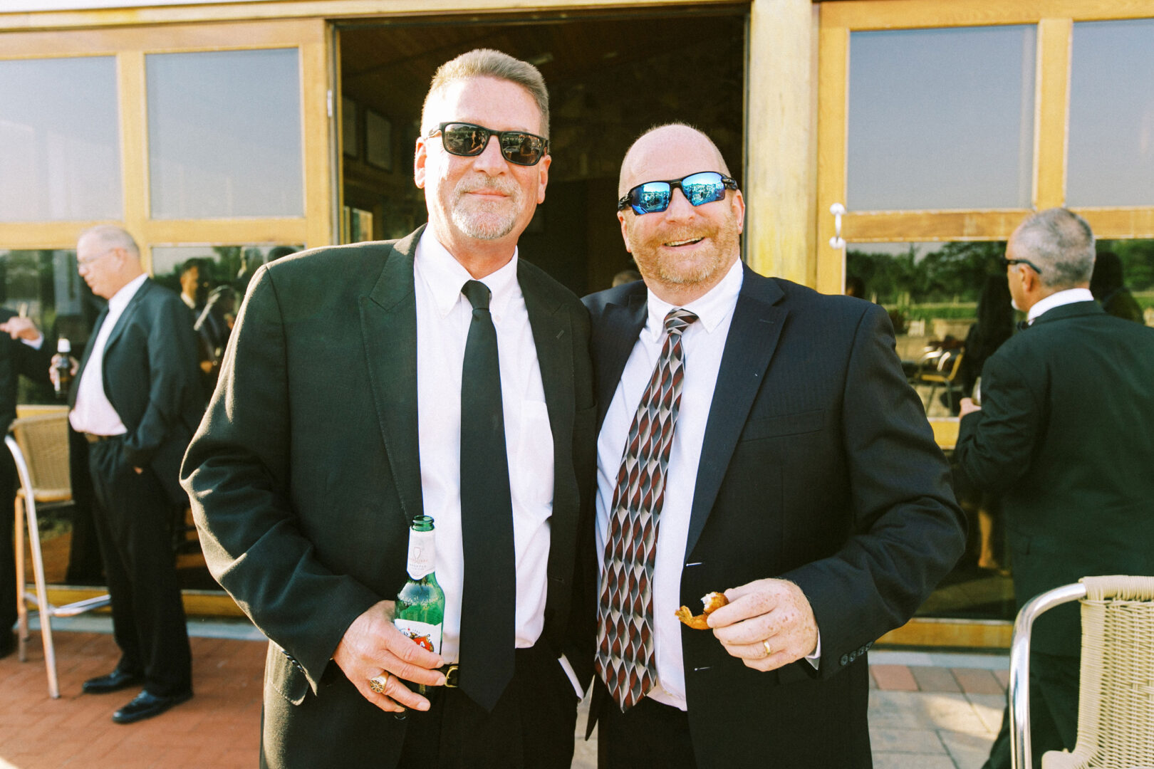 Two men in suits, wearing sunglasses, stand together outdoors at a Willow Creek Winery wedding. One holds a beverage bottle, and the other has food. People in similar attire mingle in the background.