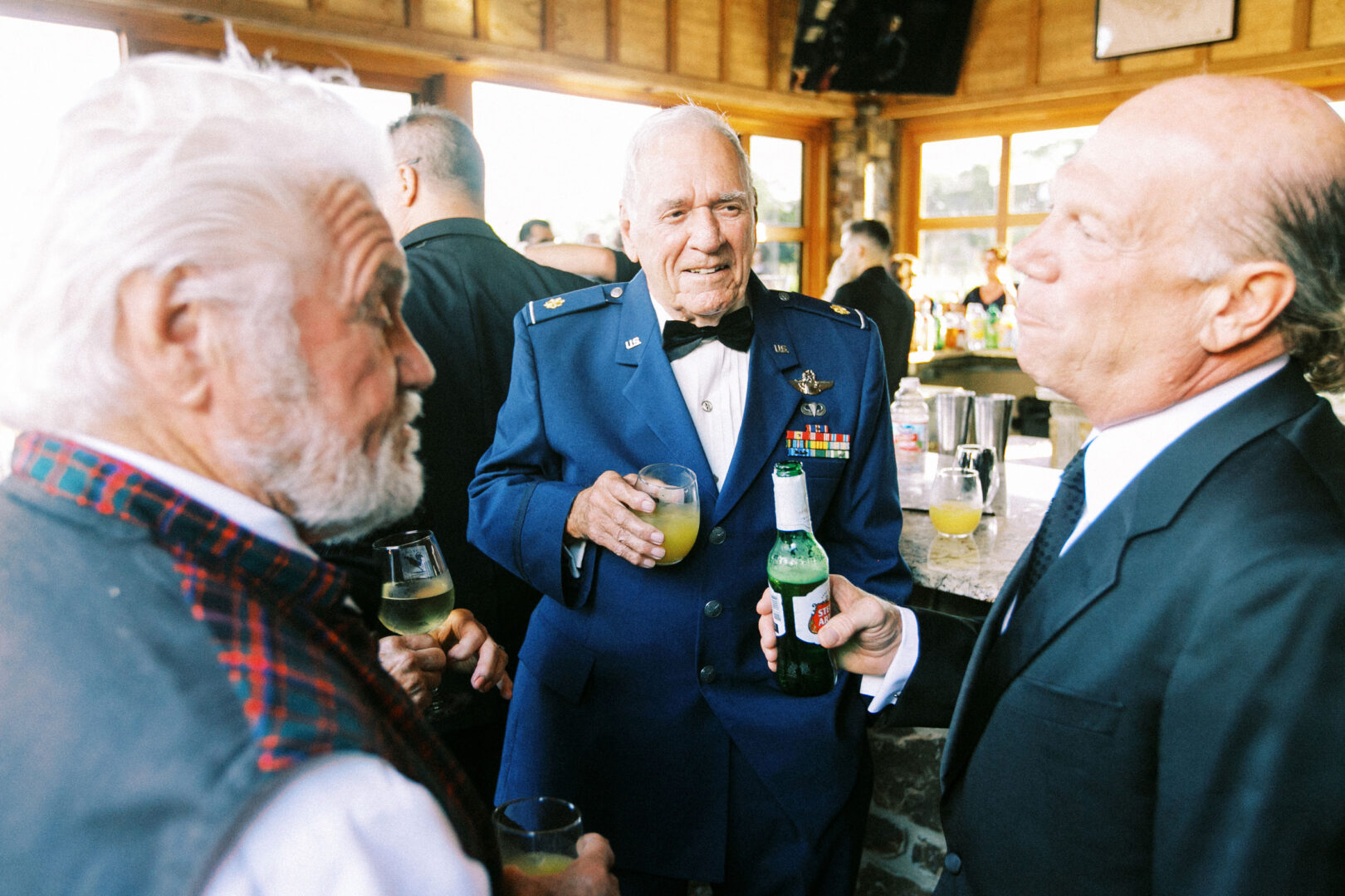 Three older men in formal attire, one in a military uniform, engage in conversation at an elegant Willow Creek Winery wedding reception, each holding a drink amidst the buzzing indoor gathering.