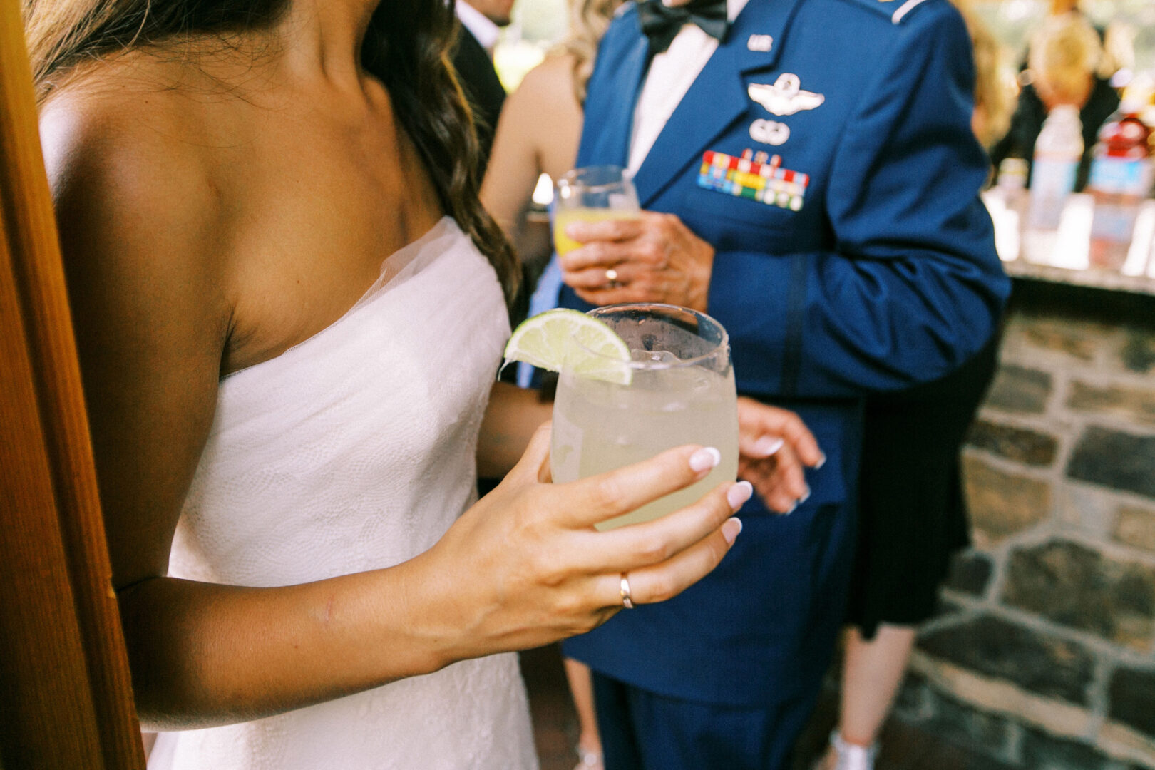 At a Willow Creek Winery wedding, a person in a strapless white dress holds a drink with a lime wedge. In the background, another person in a blue uniform enjoys a similar drink, setting the scene for an elegant celebration.