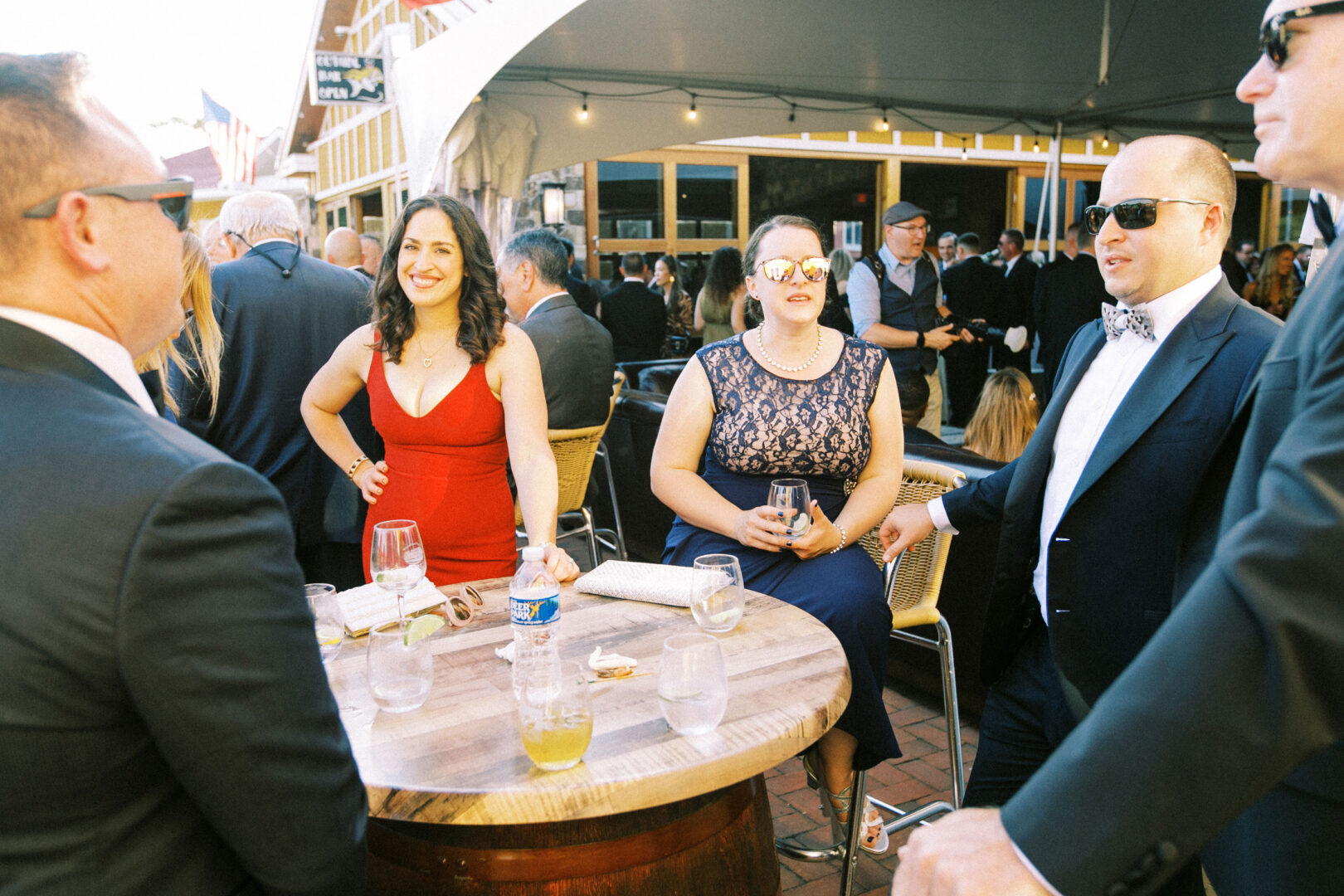 A group of people in formal attire gathers around a round table outdoors at Willow Creek Winery. Drinks and a water bottle are on the table, and some are wearing sunglasses, adding to the relaxed yet elegant wedding atmosphere.