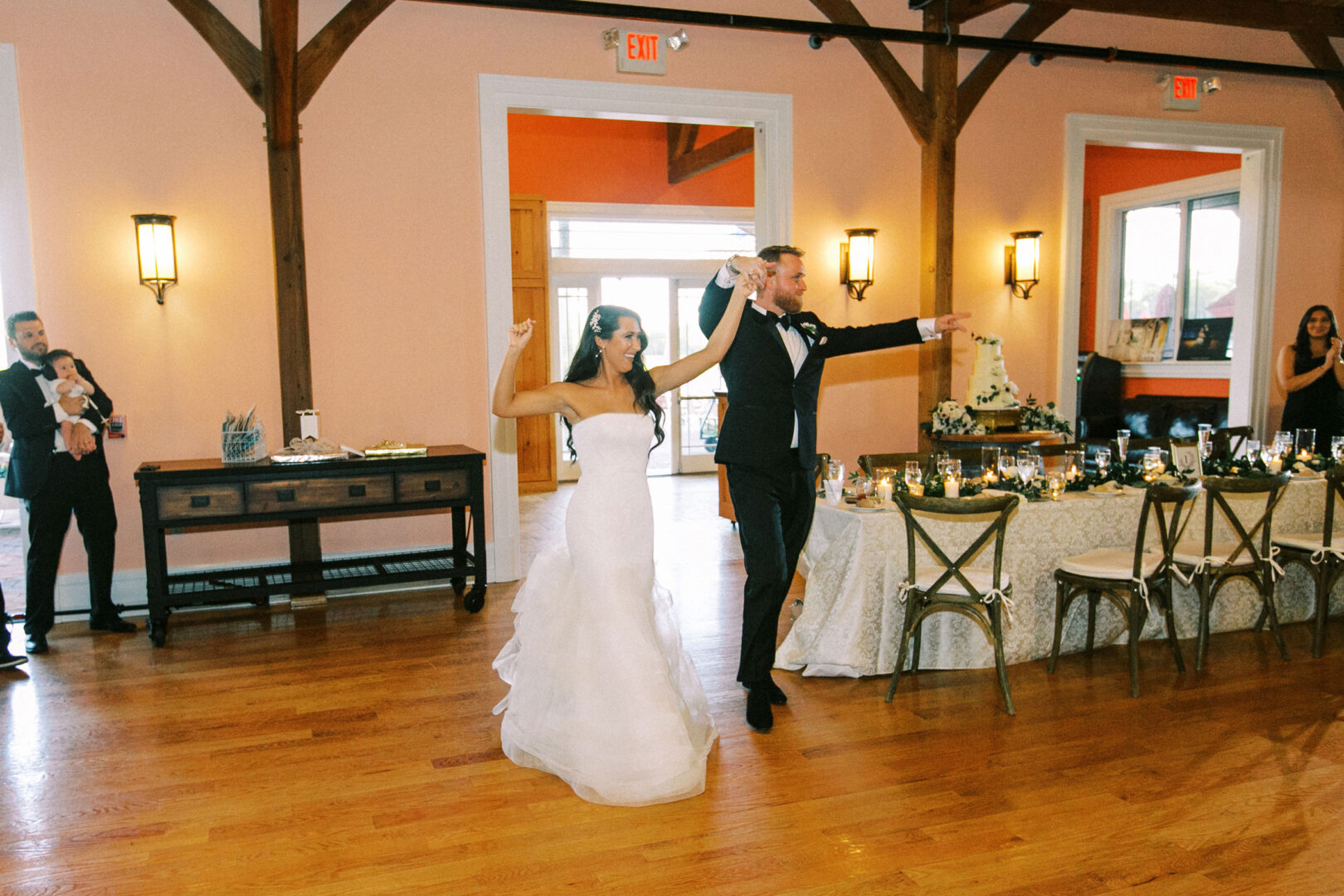 At the Willow Creek Winery wedding, a bride and groom enter the reception hall, smiling and raising their arms. Guests seated at a long table watch them. The room is adorned with wooden beams and peach-colored walls, creating an inviting and warm atmosphere.