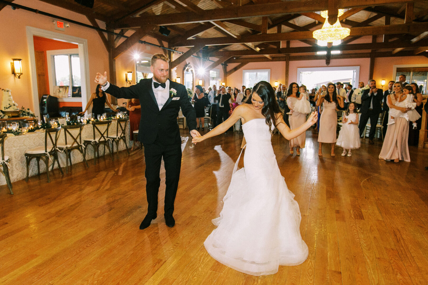 Amidst the rustic charm of Willow Creek Winery, a bride and groom share a dance in a wooden hall surrounded by guests. She twirls gracefully in her white gown, while he looks dapper in his black suit.