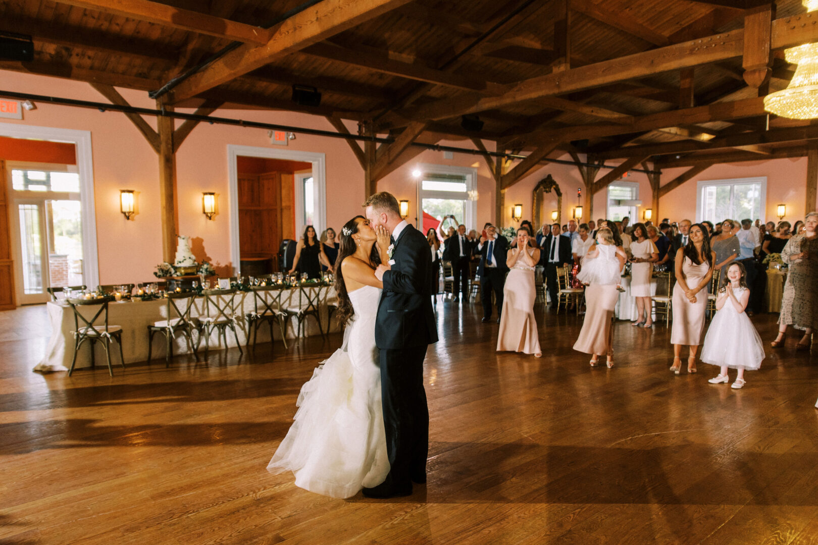 A couple shares their first dance at a Willow Creek Winery wedding reception, while guests watch and the bridal party stands nearby in a warmly lit hall.