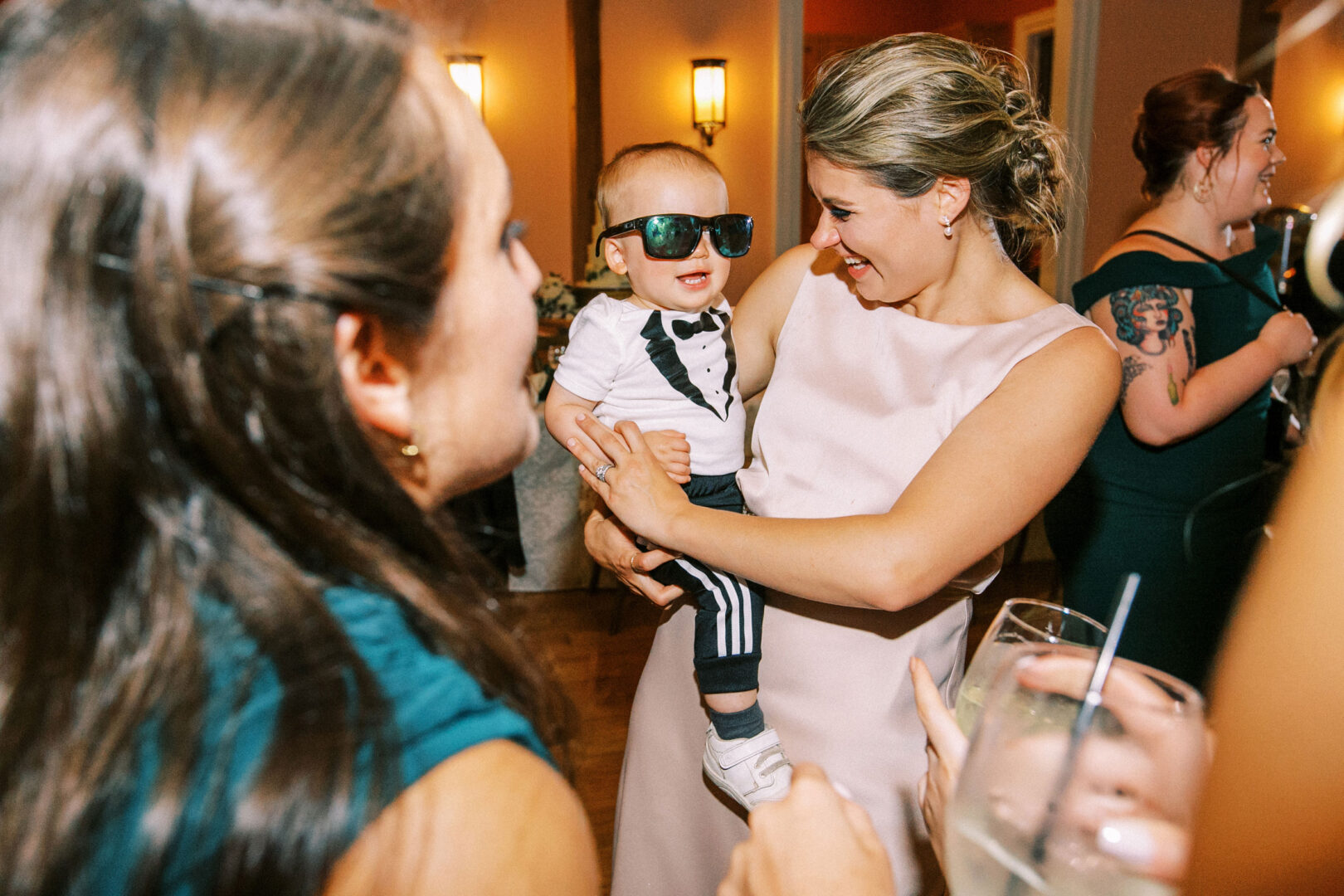 At a charming Willow Creek Winery wedding, a woman in a pink dress holds a baby wearing sunglasses and a tuxedo-print shirt. Guests stand nearby with drinks, engaging in lively conversation.