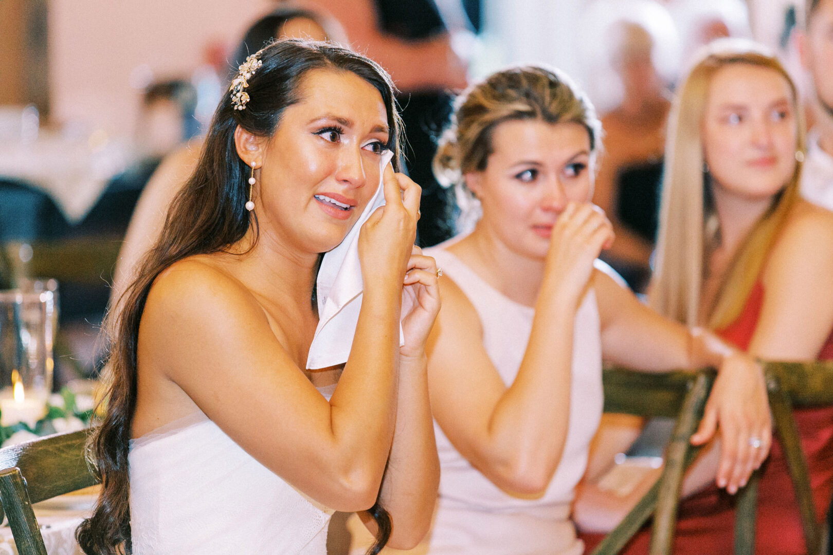 At a Willow Creek Winery wedding, two women are seated, visibly emotional, and wiping tears with tissues. Other people are seen in the background, adding to the heartfelt scene amid rows of lush vines and romantic charm.